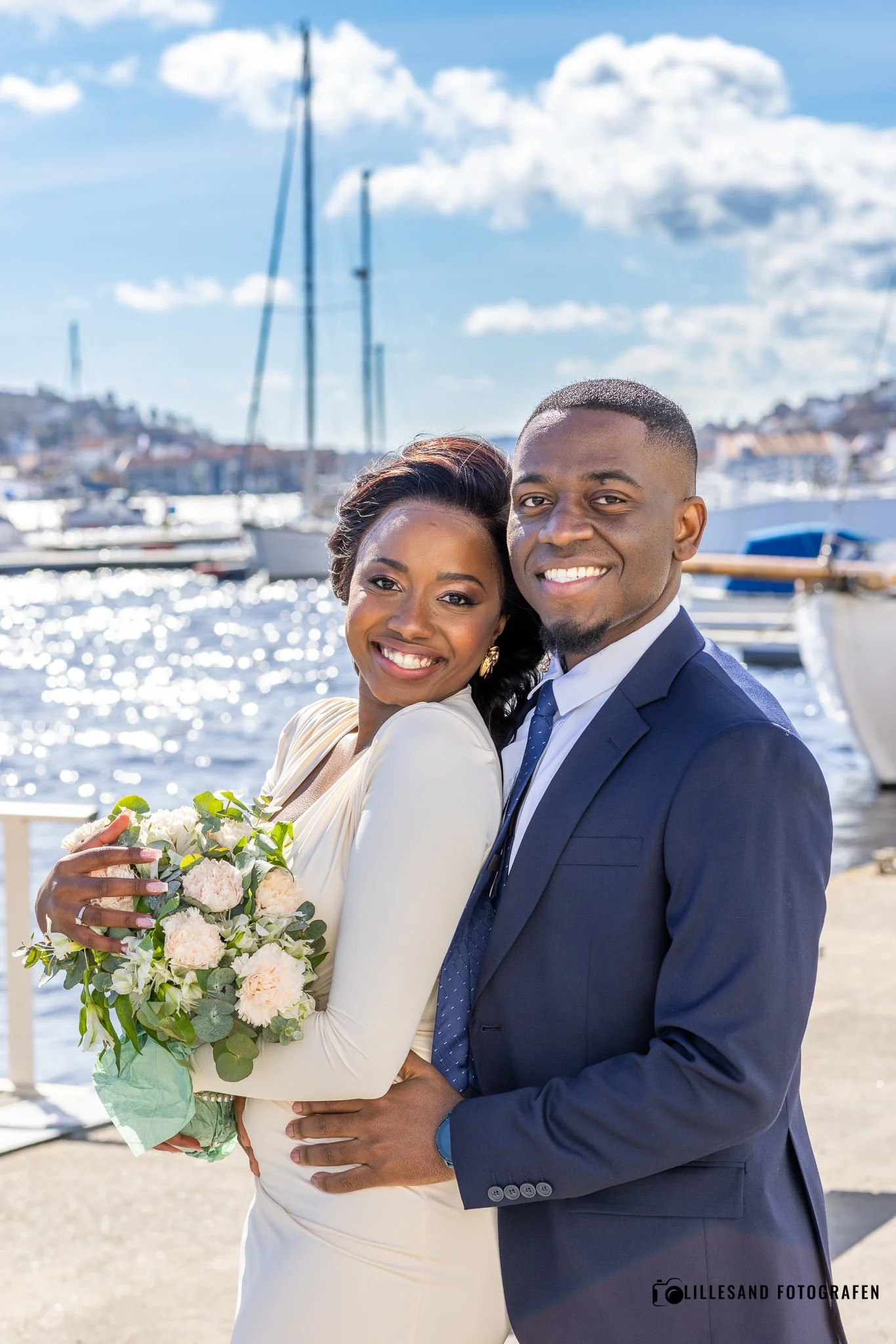 En lykkelig brudepar står ved marina, kvinnen holder en blomsterbukett, begge smiler mot kamera, med båter i bakgrunnen under blå himmel.