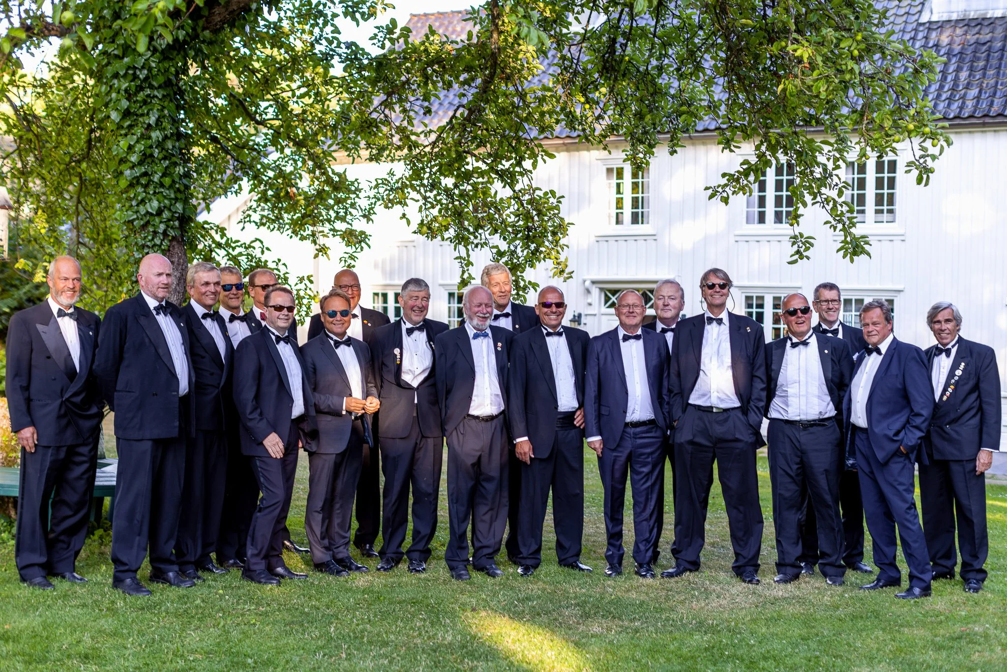 En gruppe menn i smoking står sammen utendørs under et tre, foran en hvit bygning.