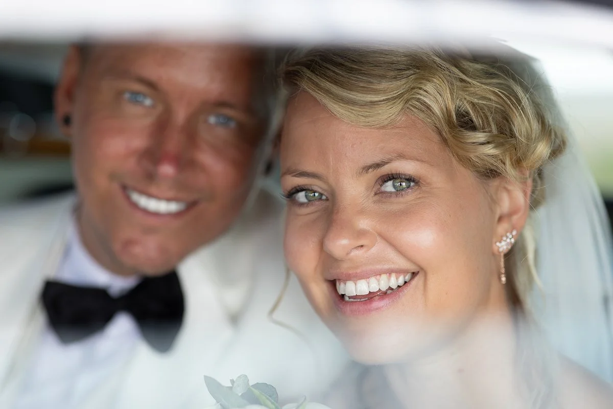 Et lykkelig brudepar smiler i en bil, med den brudens ansikt tydelig i forgrunnen og den grooms ansikt delvis synlig i bakgrunnen.