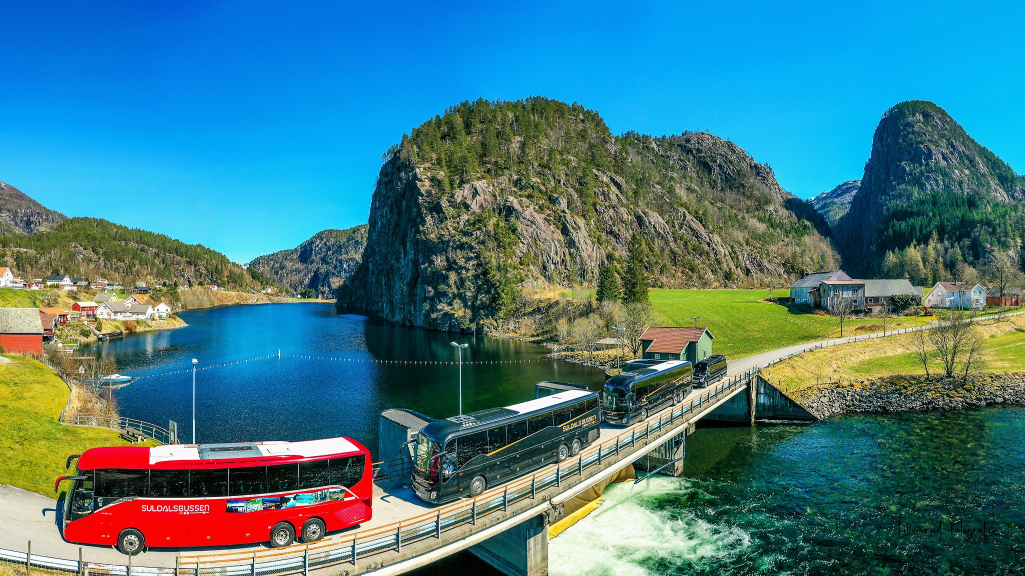 En rød buss og to svarte busser kjører over en bro over en sjø, omgitt av grønne åser og fjell med små hus, under en klar blå himmel.