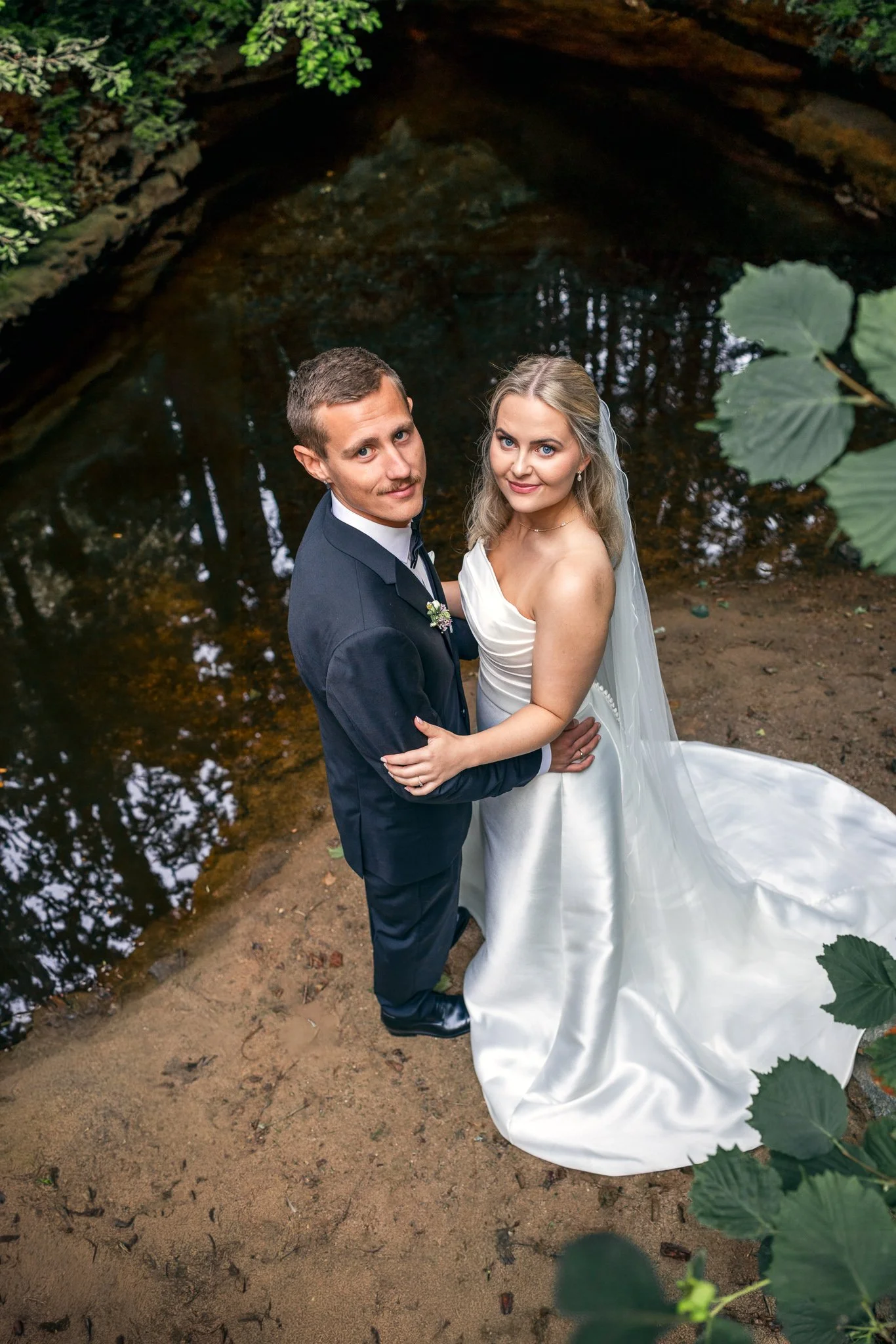 Et bilde av et kledd brudepar som står nær en skogbekk, sett ovenfra. Bruden har langt, blond hår og hvit kjole, mens brudgommen har kort, brunt hår og mørk dress.