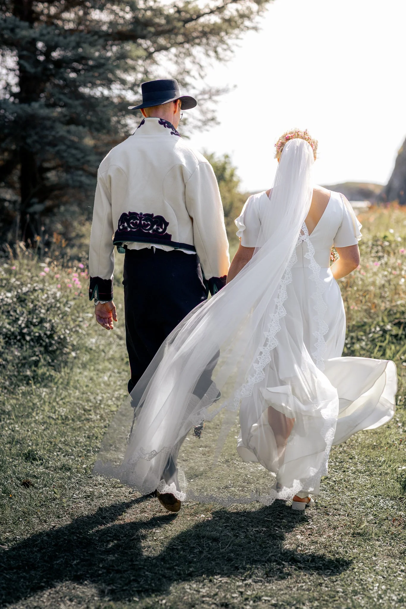 To personer går utendørs, en kvinne i en hvit brudekjole og en mann i en lys jakke og hatt.