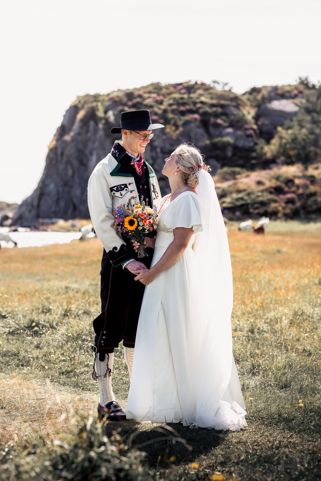 Et par i bryllupskledning står på en eng med fjell i bakgrunnen, de ser på hverandre og holder hender. Den mannlige er iført tradisjonelle norske bunader ogholder en blomsterbukett.