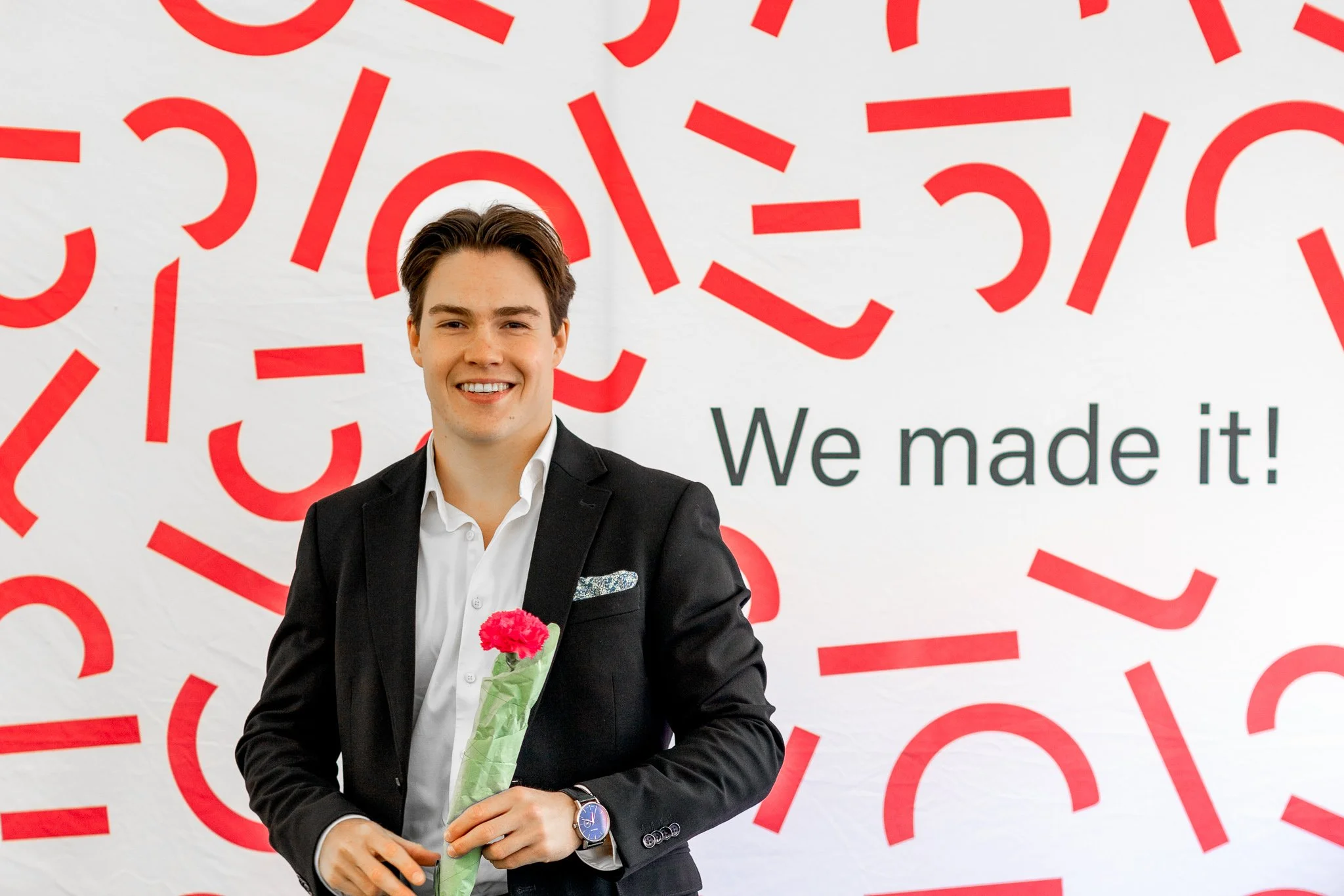 En mann i dress med hvit skjorte holder en rosa blomst, står foran en bakgrunn med røde mønstre og teksten 'We made it!'