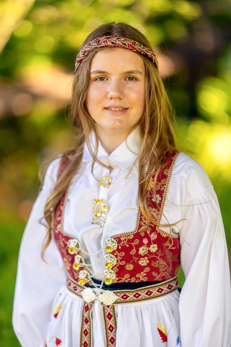 Ung jente i tradisjonell norsk bunad med blomsterprint, gullkjede og broderi, utendørs med grønn bakgrunn.
