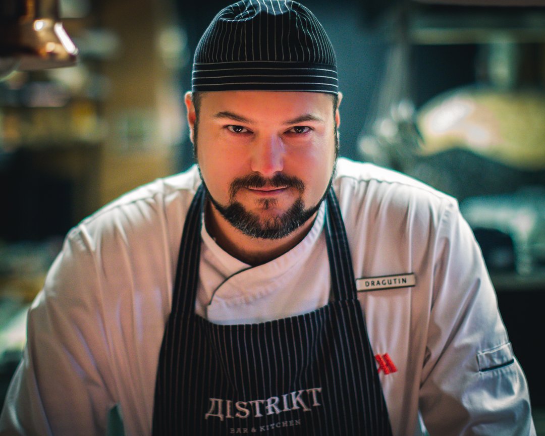 En mann i kokkeuniform med svart og hvit stripete kokkehat, svart og hvit stripete forkle med teksten "Distrikt Bar & Kitchen". Han har et navnmerke med navnet "Dragutin" og ser direkte mot kamera med et smil.