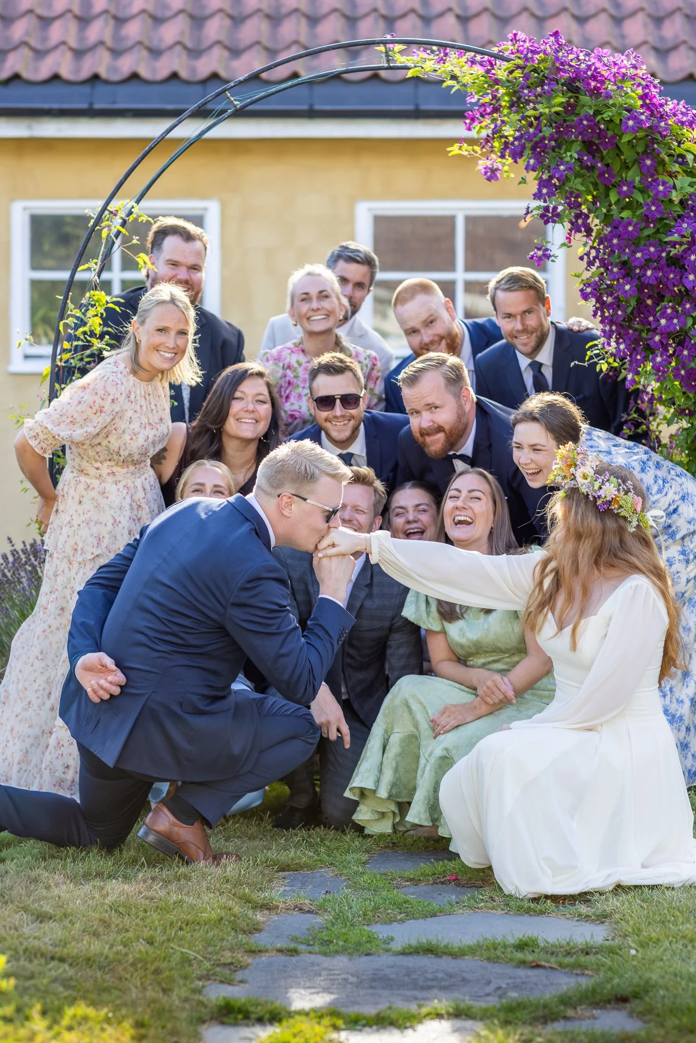 En gruppe mennesker som har et bryllup, hvor noen av dem er i festklær, og noen er i brudekjoler og dress, som står bak en romantisk port dekket med lilla blomster.