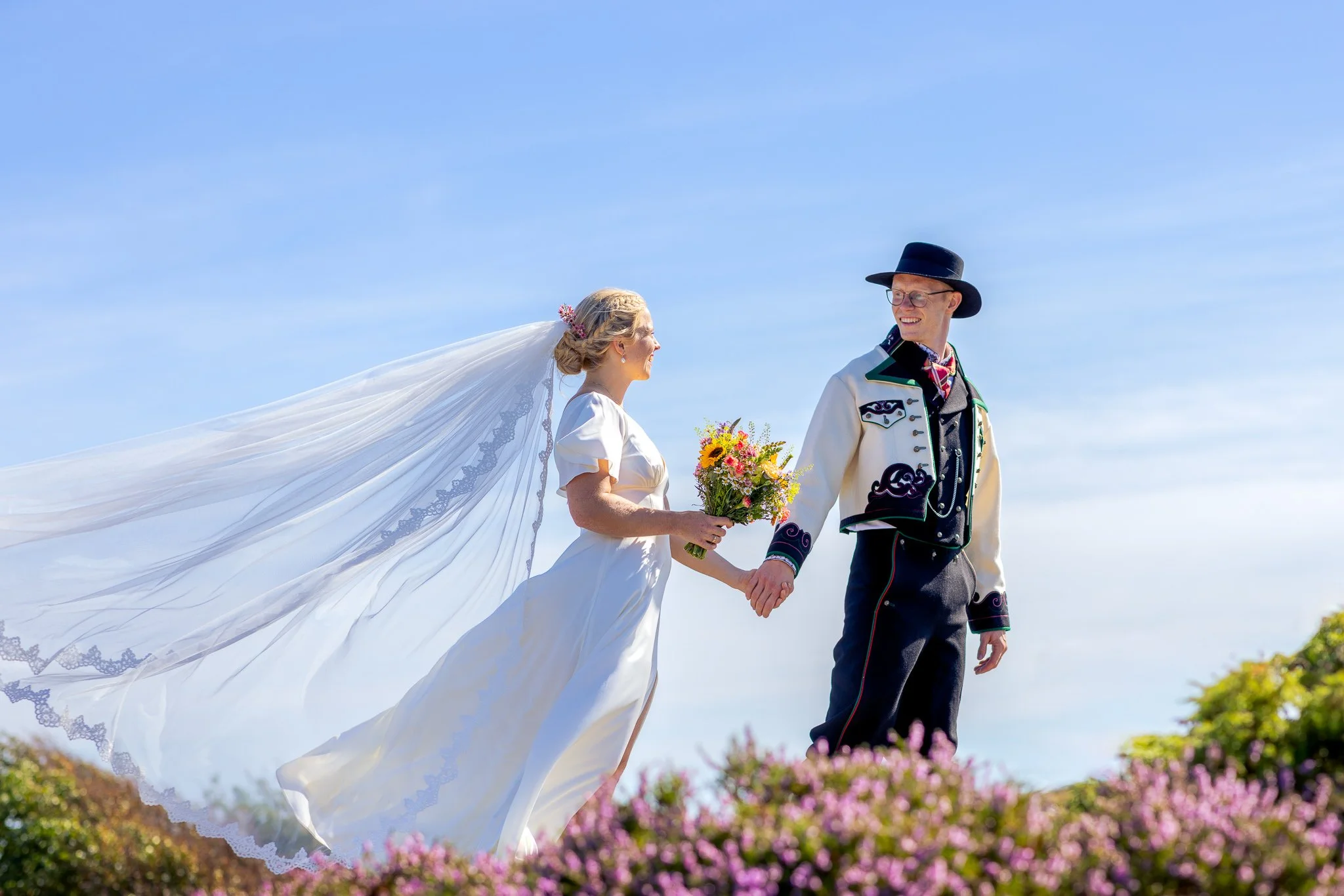 En brud og en brudgom holder hender utendørs med blomster i hånden, under en blå himmel, iført tradisjonelle klede.