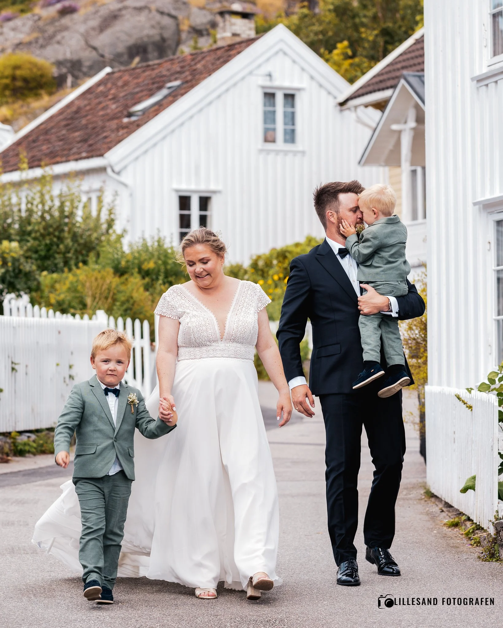 En bryllupsfamilie går nedover en gate, med hus i bakgrunnen. Kvinne i hvit brudekjole, mann i dress med barn, og en liten gutt i dress holder hender.