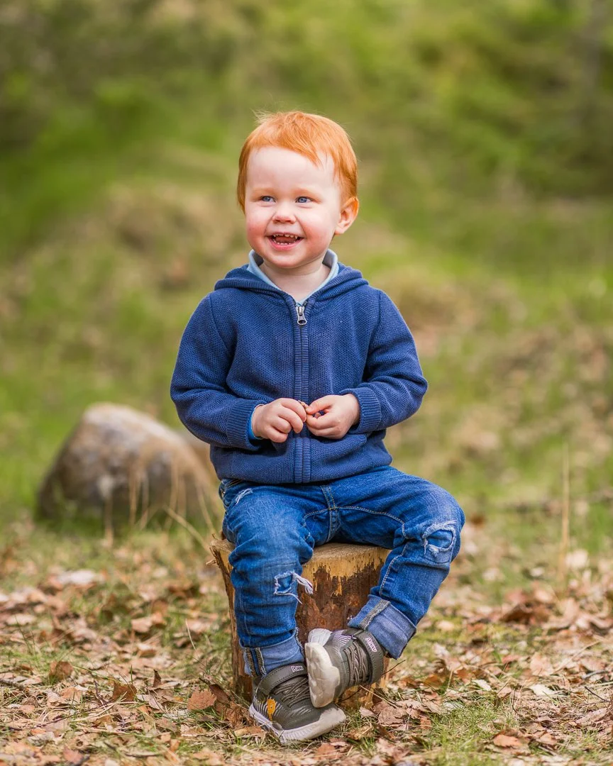 Ung gutt med rødt hår smiler mens han sitter på en stubbe i en skog med grønne blader og fallne blad på bakken.