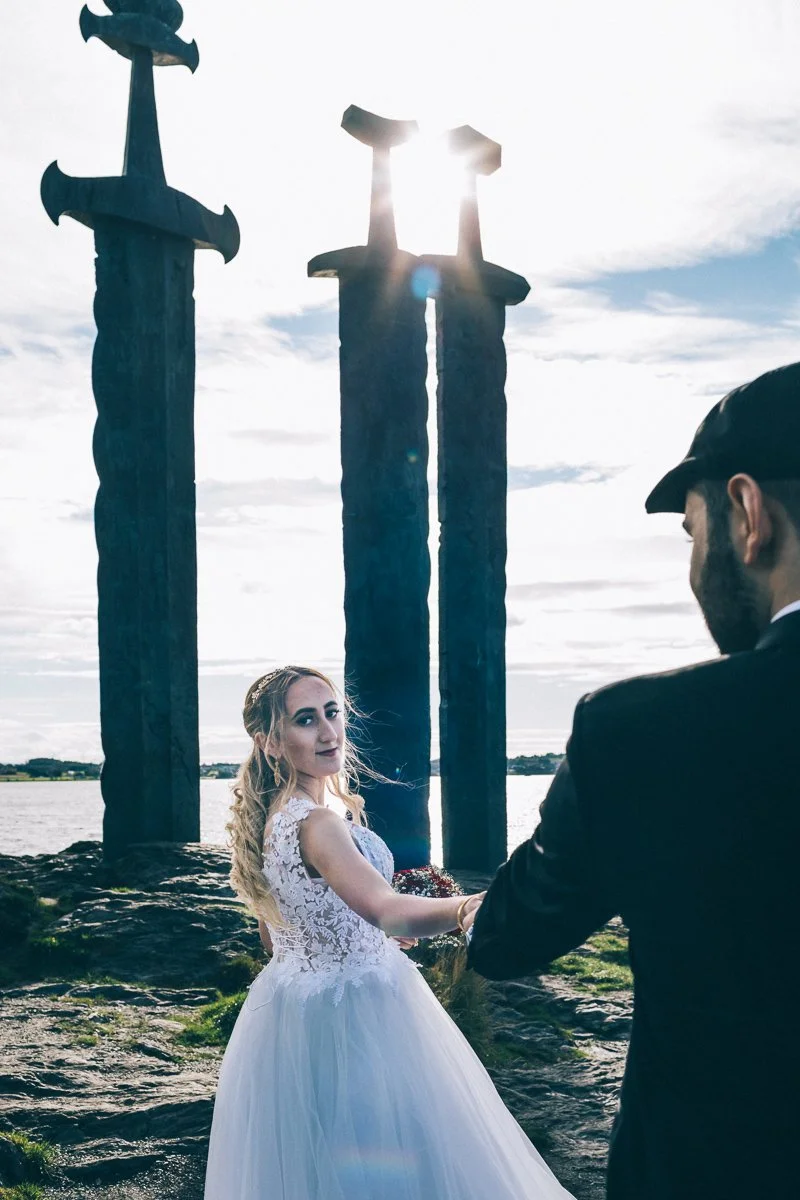 En brud og brudgom holder hender foran tre store sverd-figurer på stranden, med vann og en skyet himmel i bakgrunnen.