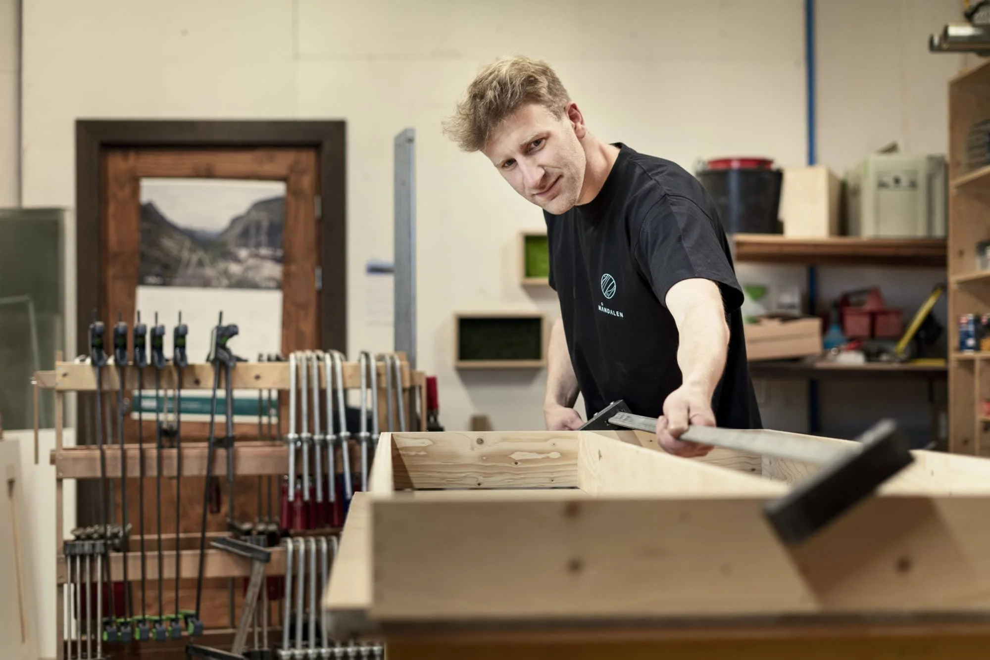 En mann arbeider med tre i et verksted, brukende et mål og en bormaskin. Verkstedet har hyller med utstyr.