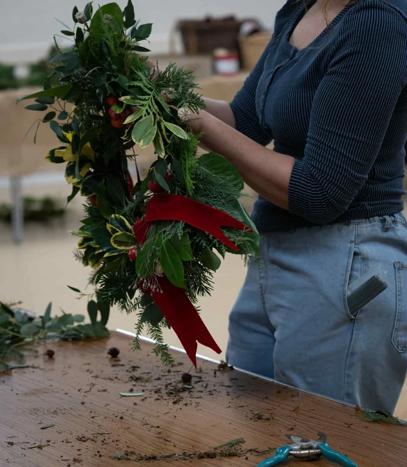 Person assembling a festive holiday wreath with greenery and red accents on a wooden table.