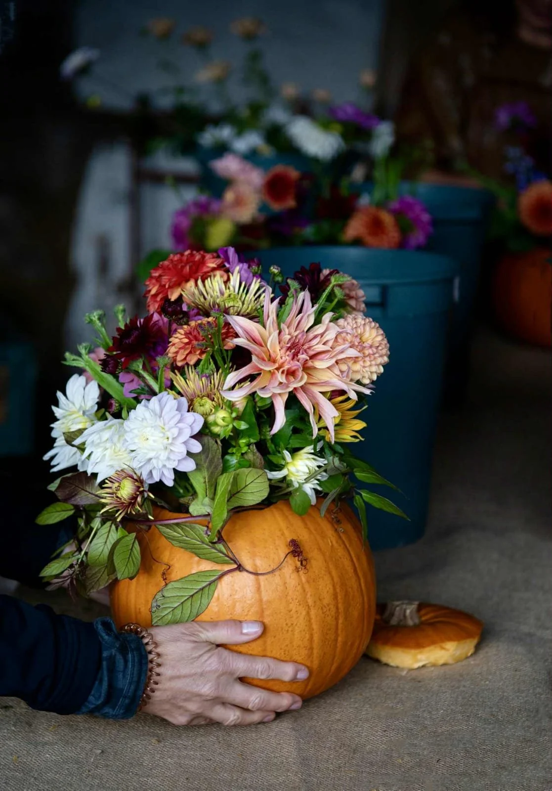 A handmade floral arrangement in the shape of a pumpkin, with pink, white, and purple flowers and green leaves, on a table with a person's hand holding the pumpkin.
