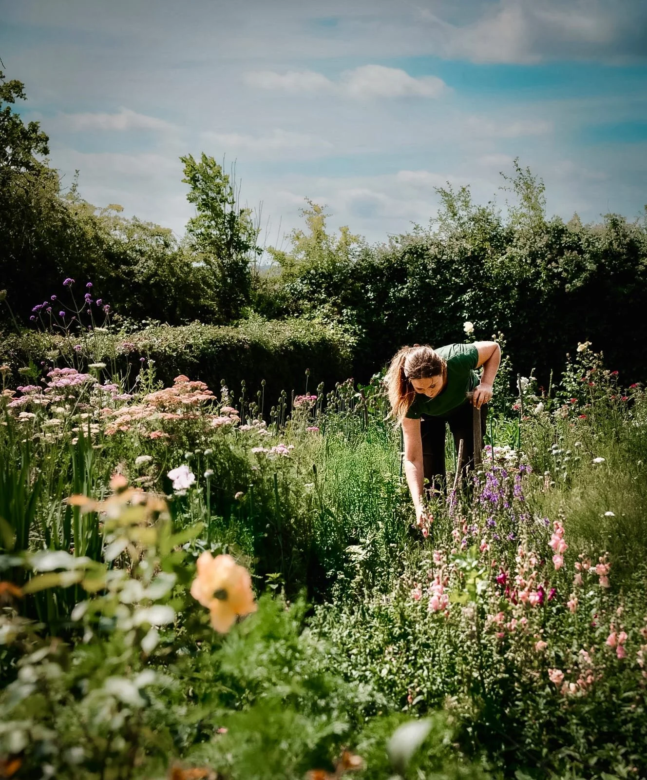 A woman with long hair bending over and touching flowers in a lush garden filled with various colorful blooming flowers, with trees and a partly cloudy sky in the background.