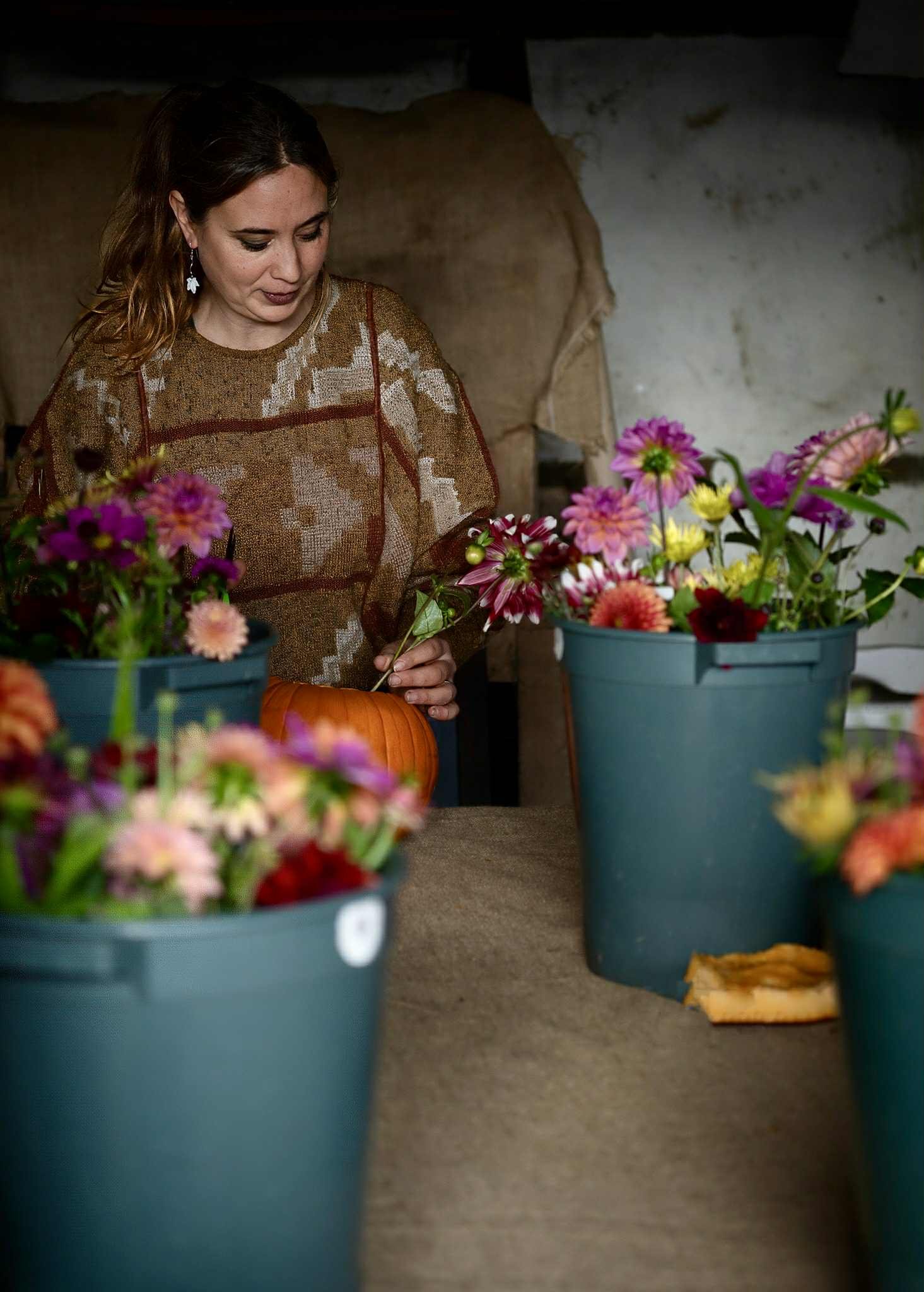 A woman with wavy brown hair, earrings, and dark lipstick, arranging flowers in a large blue pot. She is wearing a brown patterned top and is indoors, with a rough wall in the background, surrounded by similar flower arrangements.