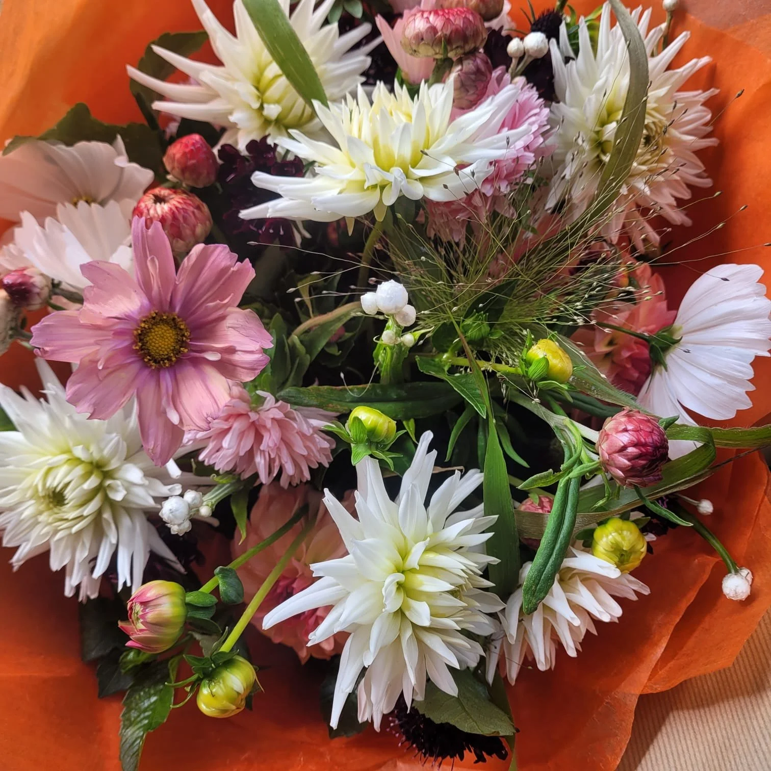 A colorful bouquet of various flowers including white, pink, and purple blooms, wrapped in orange paper.