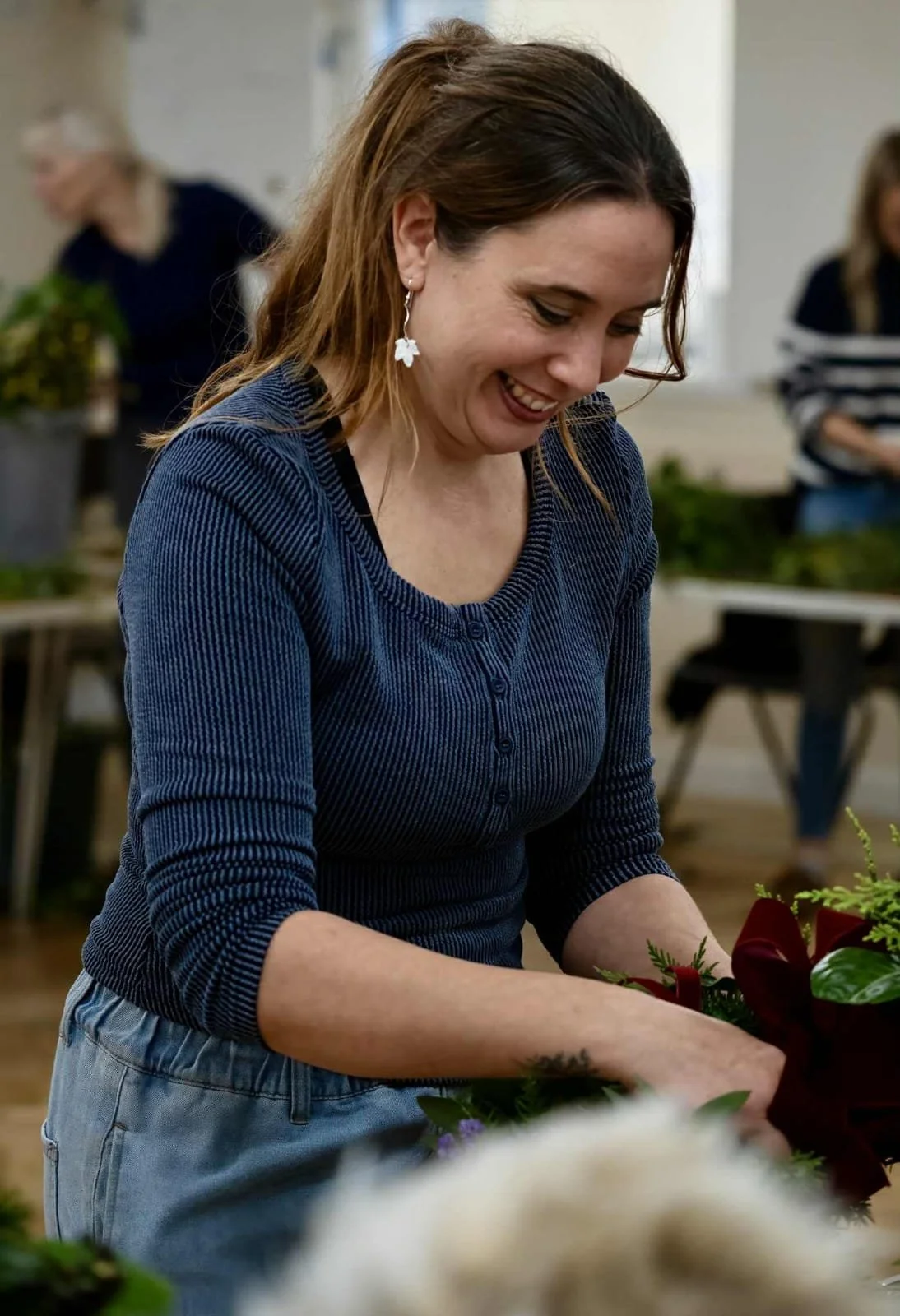 A woman with brown hair and light skin, smiling while working with plants or flowers in an indoor setting, wearing a blue ribbed long sleeve shirt and earrings with flower shapes.
