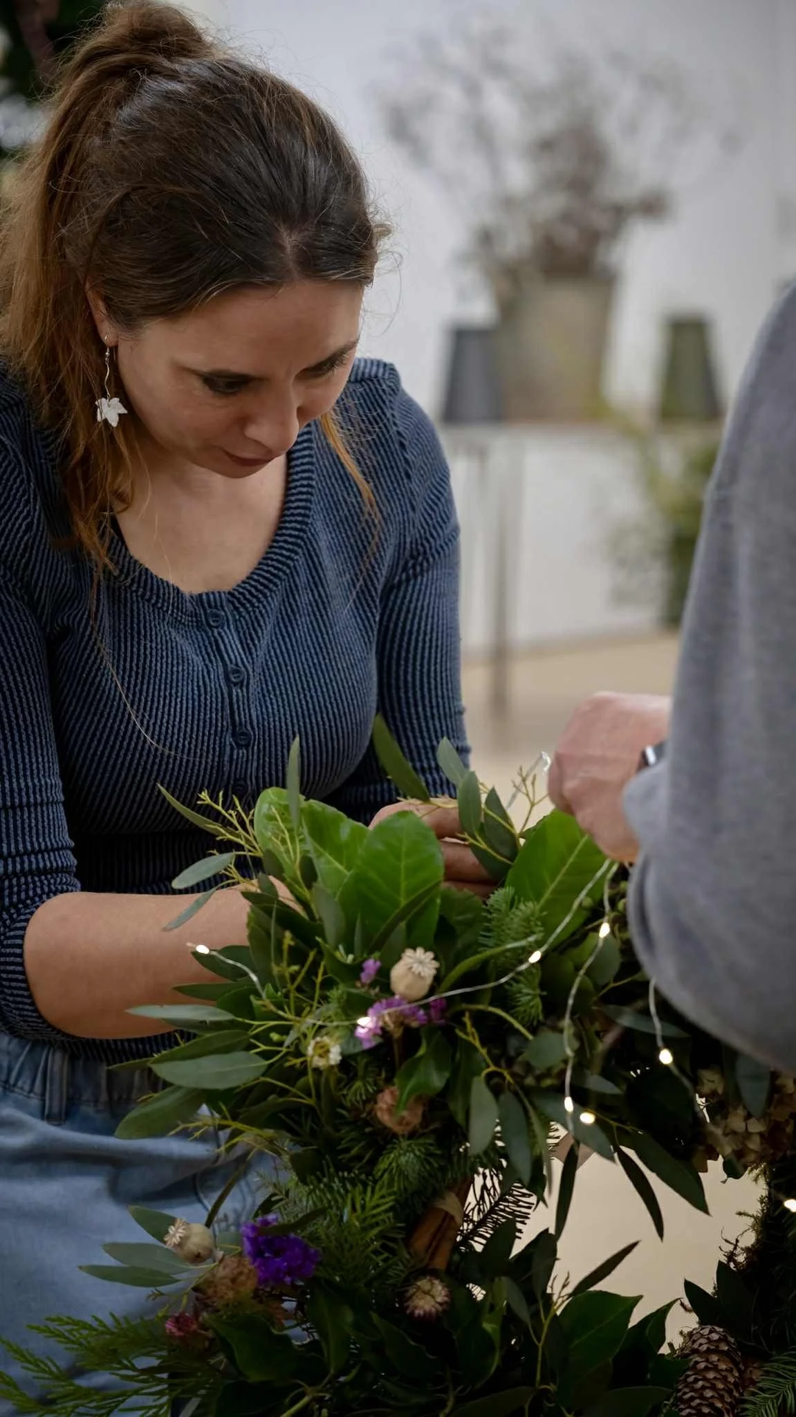 A woman is arranging a flower arrangement, looking intently at the flowers and greenery she is working with. There is an assortment of flowers and green leaves in front of her.