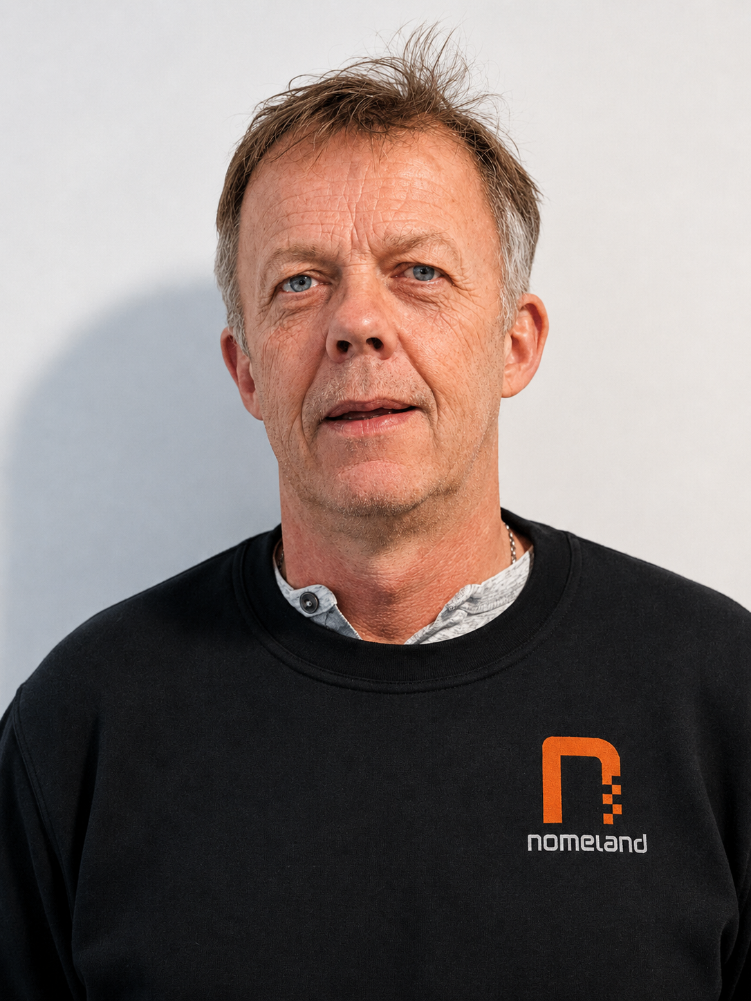 En mann med blond hår og blå øyne, iført svart genser med logoen 'nomeland' i oransje, mot en lys bakgrunn.