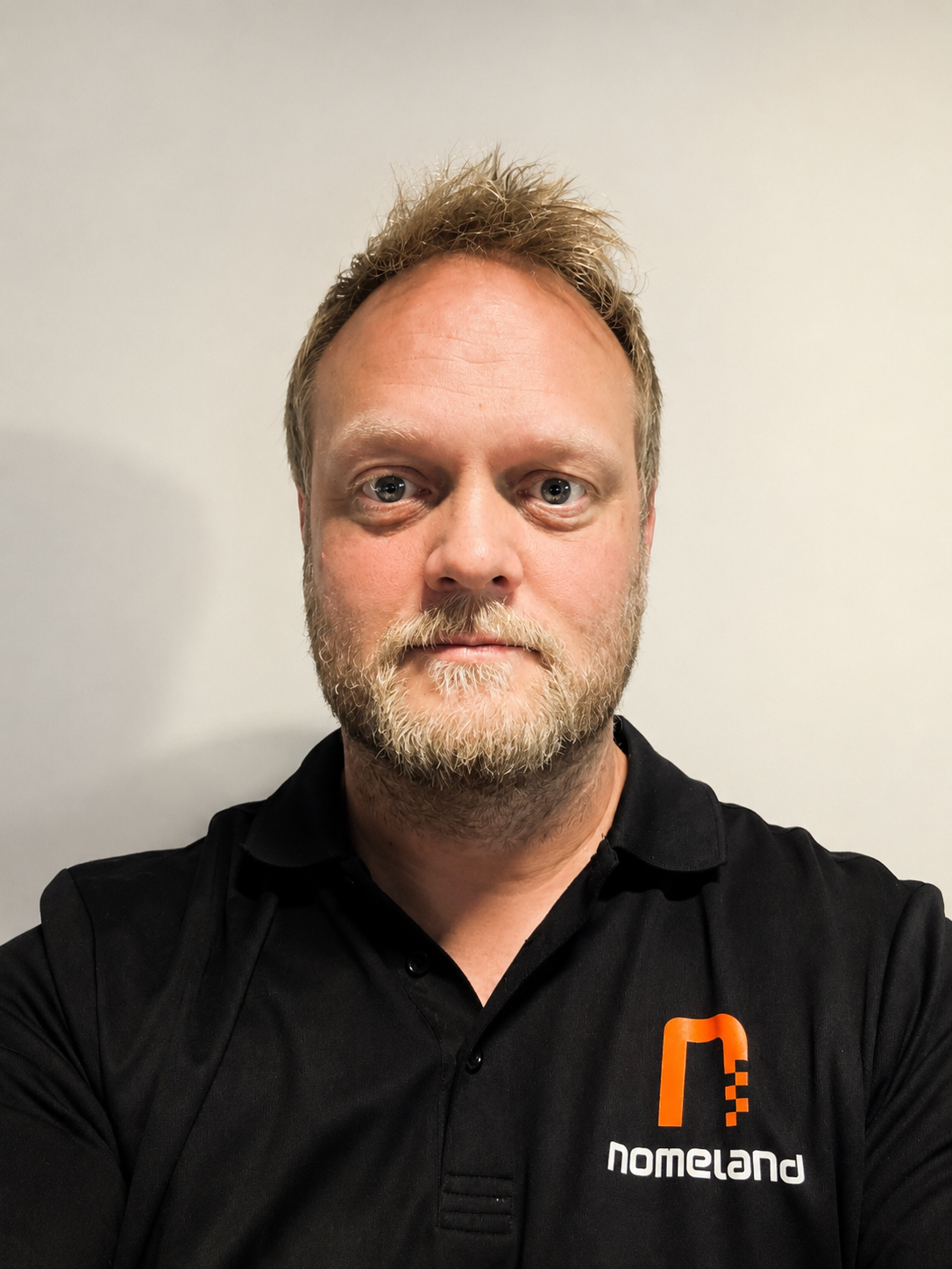 En mann med skjegg og lyst hår som tar selfie mot en hvit vegg, iført svart polo med Noregland-logo.