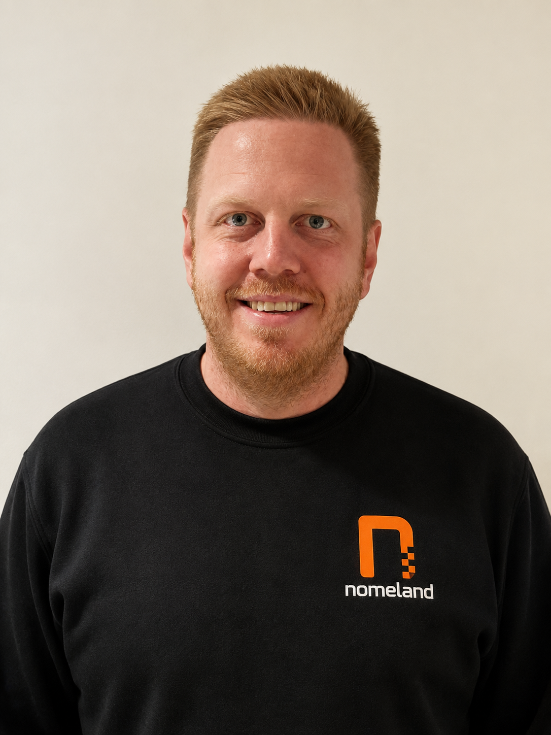 Et portrett av en mann med lys hud, rødt hår og skjegg som smiler foran en lys vegg. Han har på seg en svart genser med logoen 'nodeland' i oransje og hvitt.