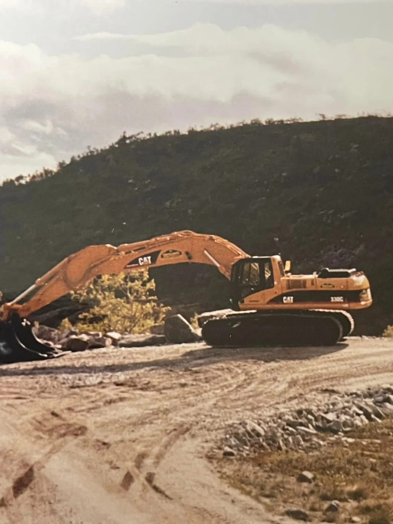 En gul excavator stående på en grusvei med et fjell i bakgrunnen under en overskyet himmel.