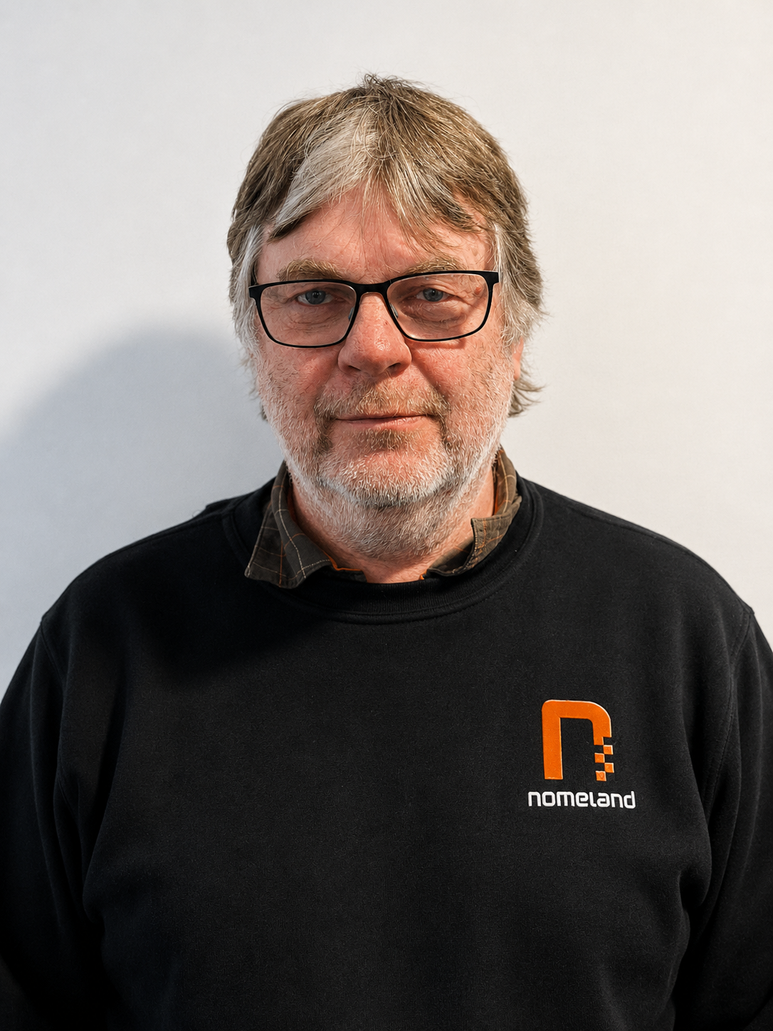 Et portrett av en eldre mann med brunt og grått hår, skjegg og briller, iført svart genser med logo og teksten 'nomeland'.