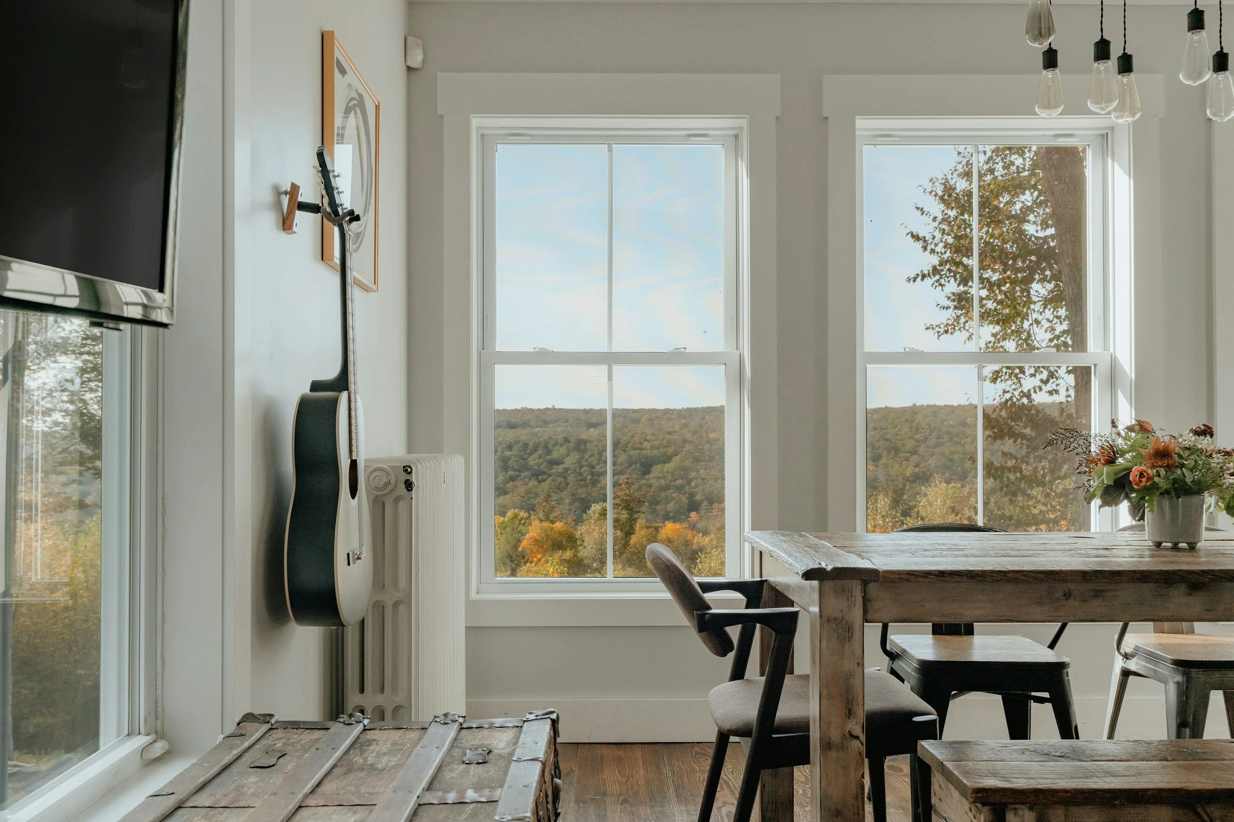 Room with large windows looking out at a landscape with hills and trees, a wooden table with a floral arrangement, a chair, vintage trunk, wall-mounted guitar, and artwork.