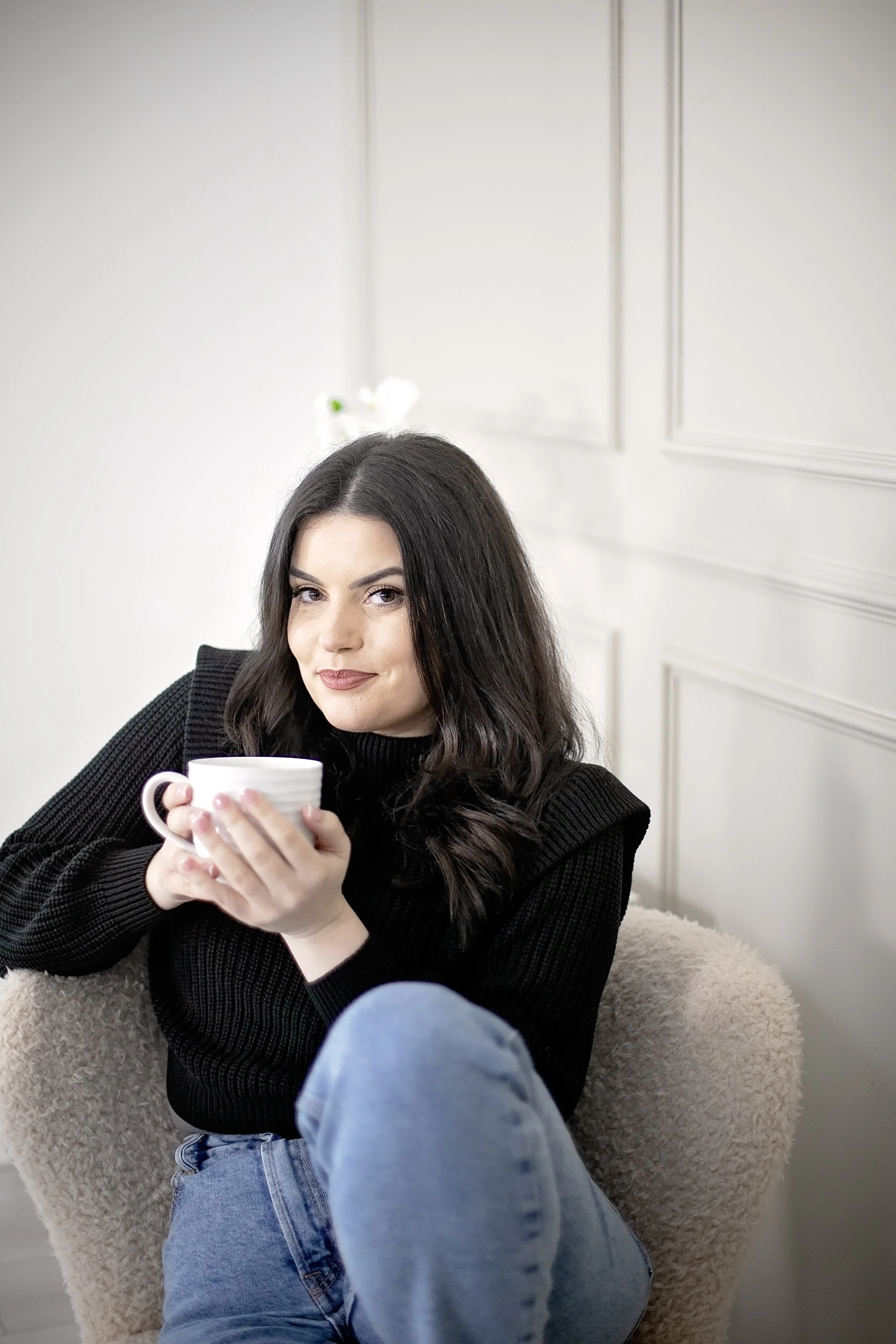 A woman with dark hair and a black sweater sitting in a cozy chair, holding a white mug and looking at the camera.