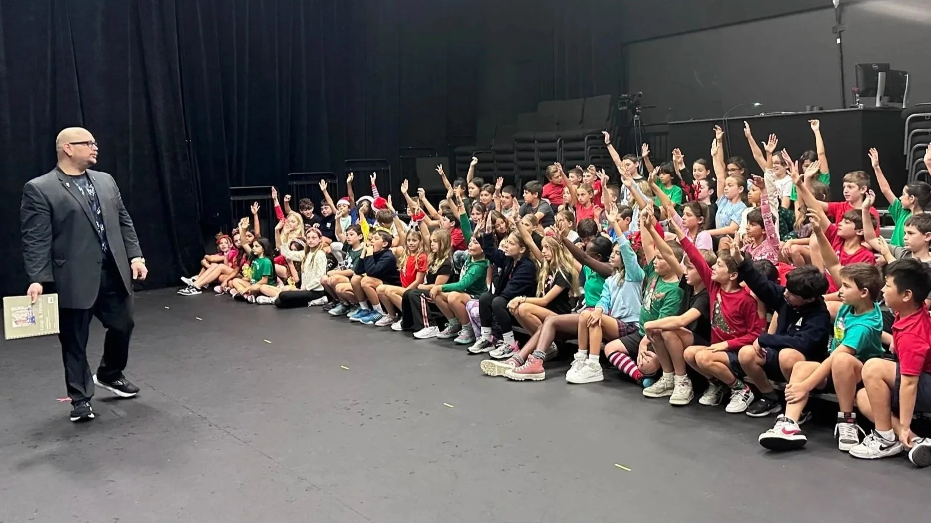 Big Joey Magic Showy professional magician asking for a volunteer in a family magic show. Children of all ages are seen raising their hands to volunteer for a magic trick.