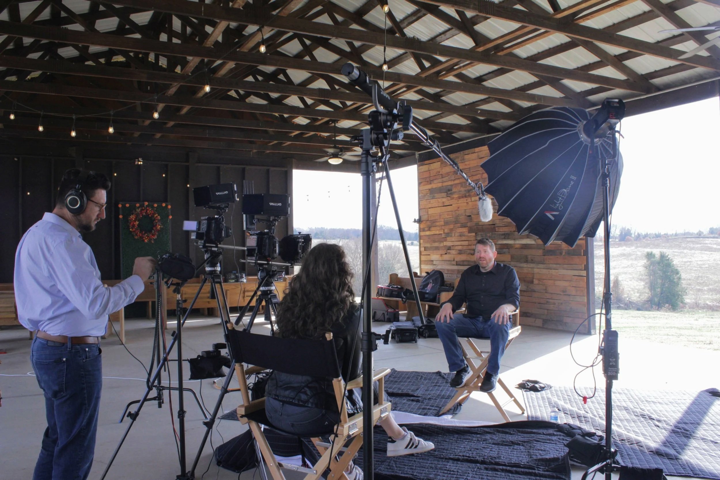 A man being interviewed on a film set with lighting and camera equipment, with a woman sitting and speaking to him, and a crew member adjusting a camera.