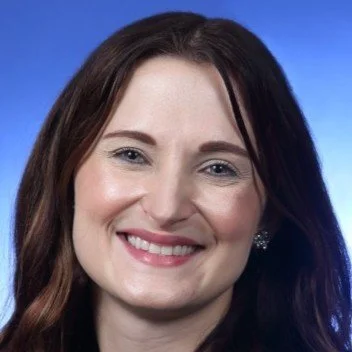 A woman with long, dark wavy hair wearing a white turtleneck sweater, smiling in a bedroom setting.