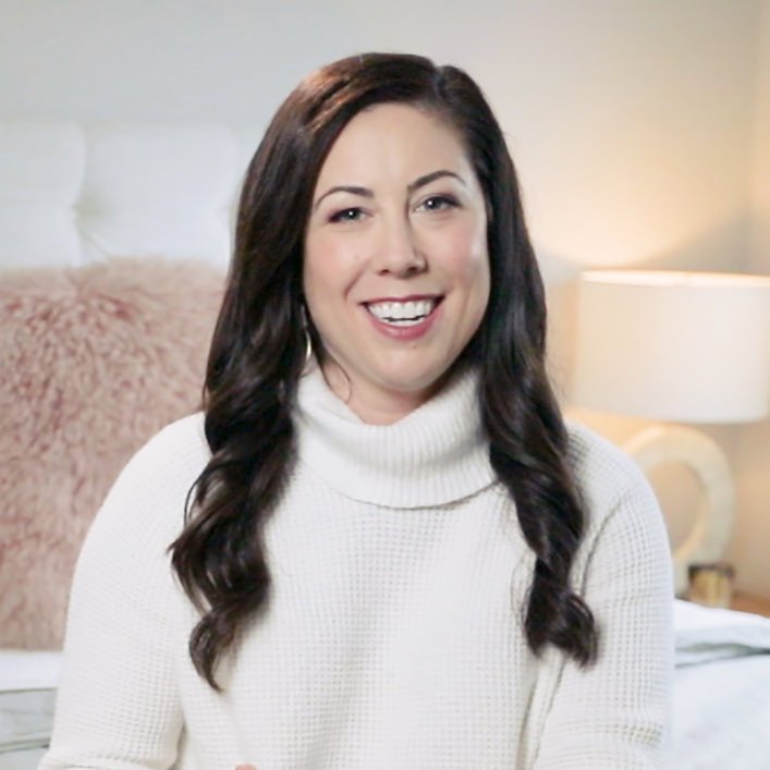 A woman with long, dark wavy hair wearing a white turtleneck sweater, smiling in a bedroom setting.