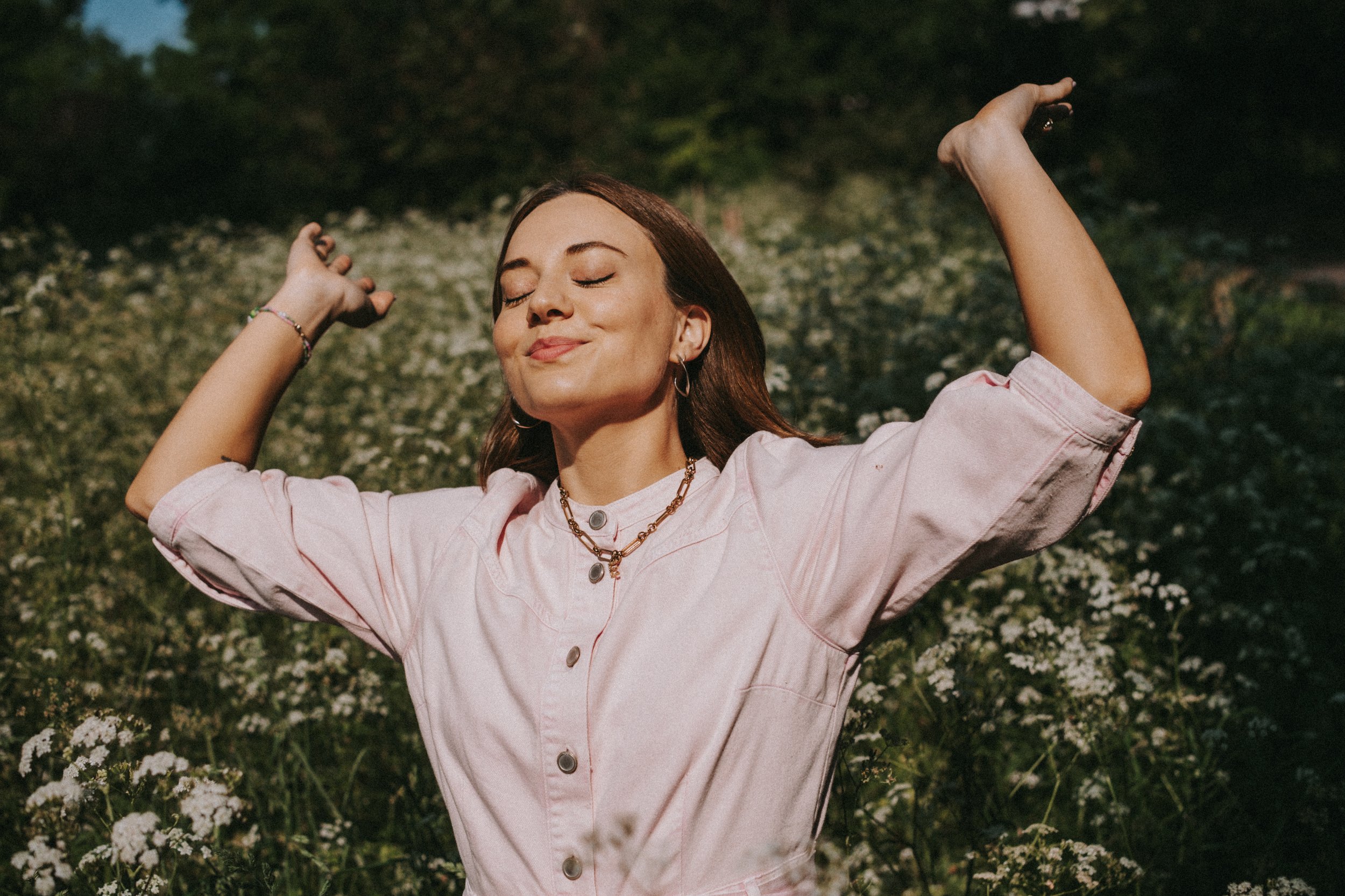 A woman with brown hair and closed eyes, smiling with arms raised, standing outdoors in a field of white flowers, wearing a light pink shirt and layered necklaces.