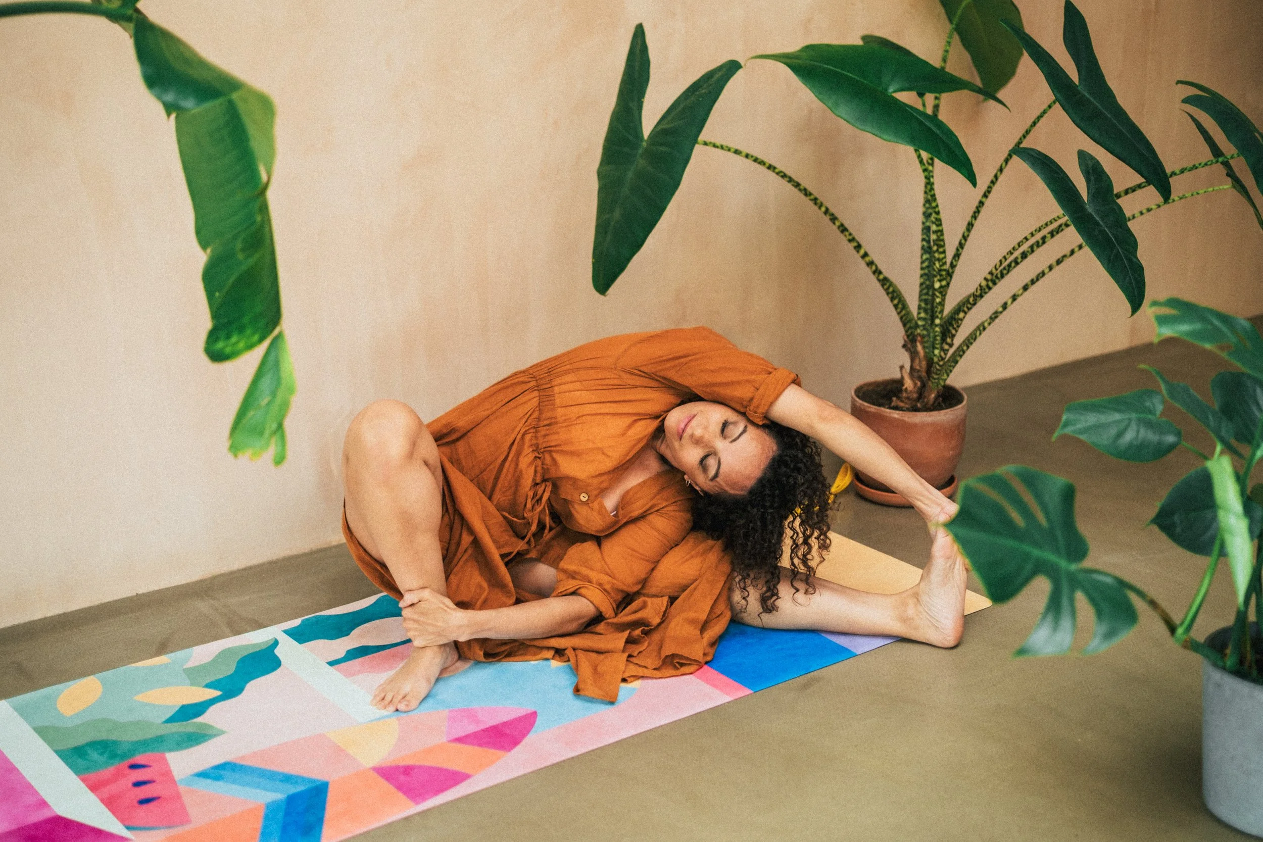 A woman practicing yoga on a colorful mat indoors, surrounded by potted green plants, wearing an orange outfit, and performing a stretch with her left arm reaching back and her right leg bent.