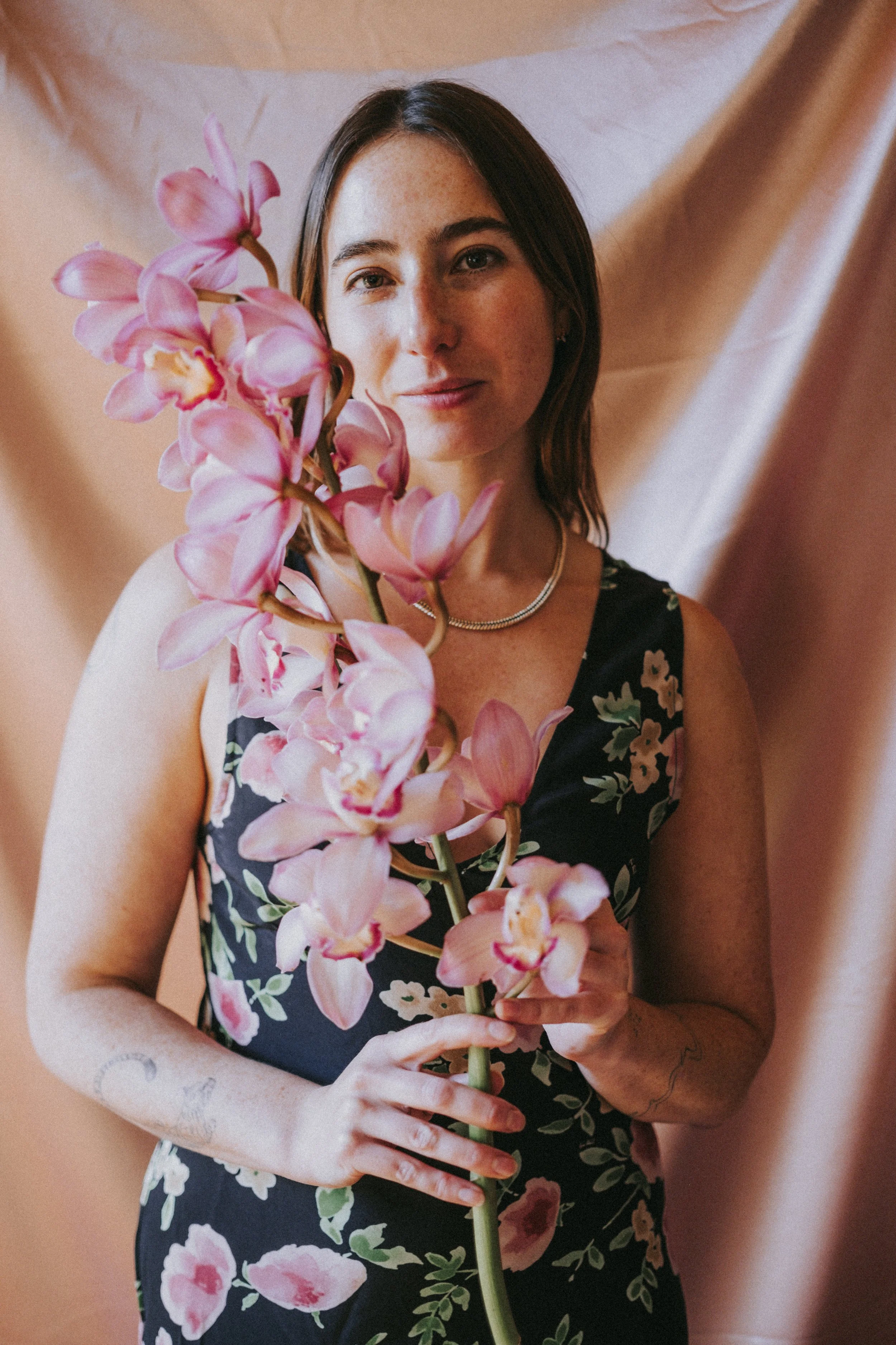 A young woman with short brown hair, wearing a black floral dress and a necklace, holds a large pink orchid flower in front of her face. She stands in front of a peach-colored backdrop.