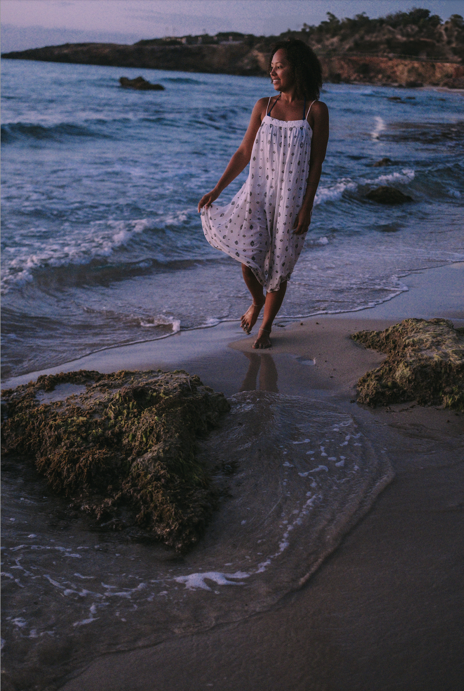 A woman in a white dress with dark polka dots walking barefoot along the shoreline at sunset, with rocks and the ocean in the background.