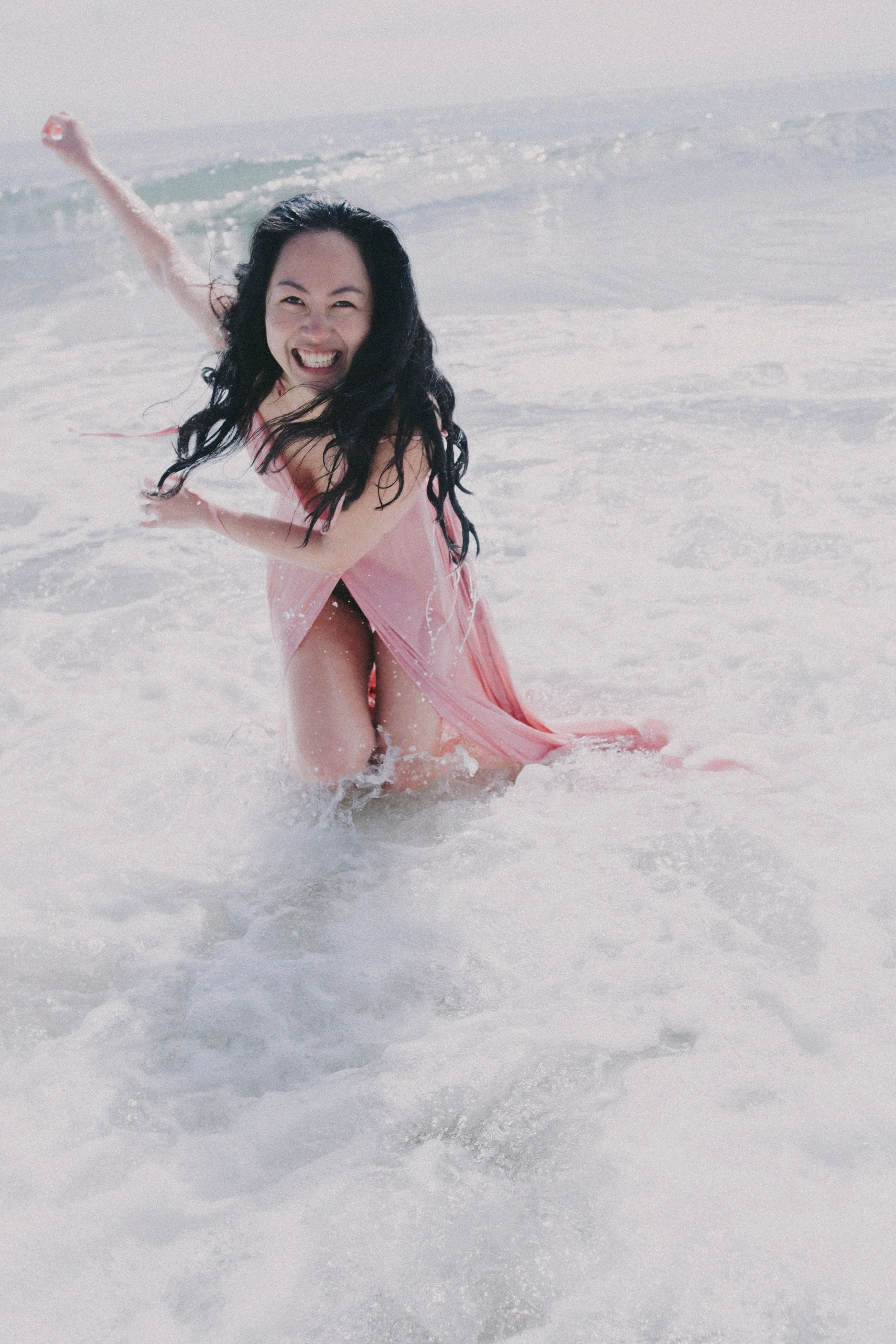 A woman in a pink dress smiling and playing in the ocean waves.