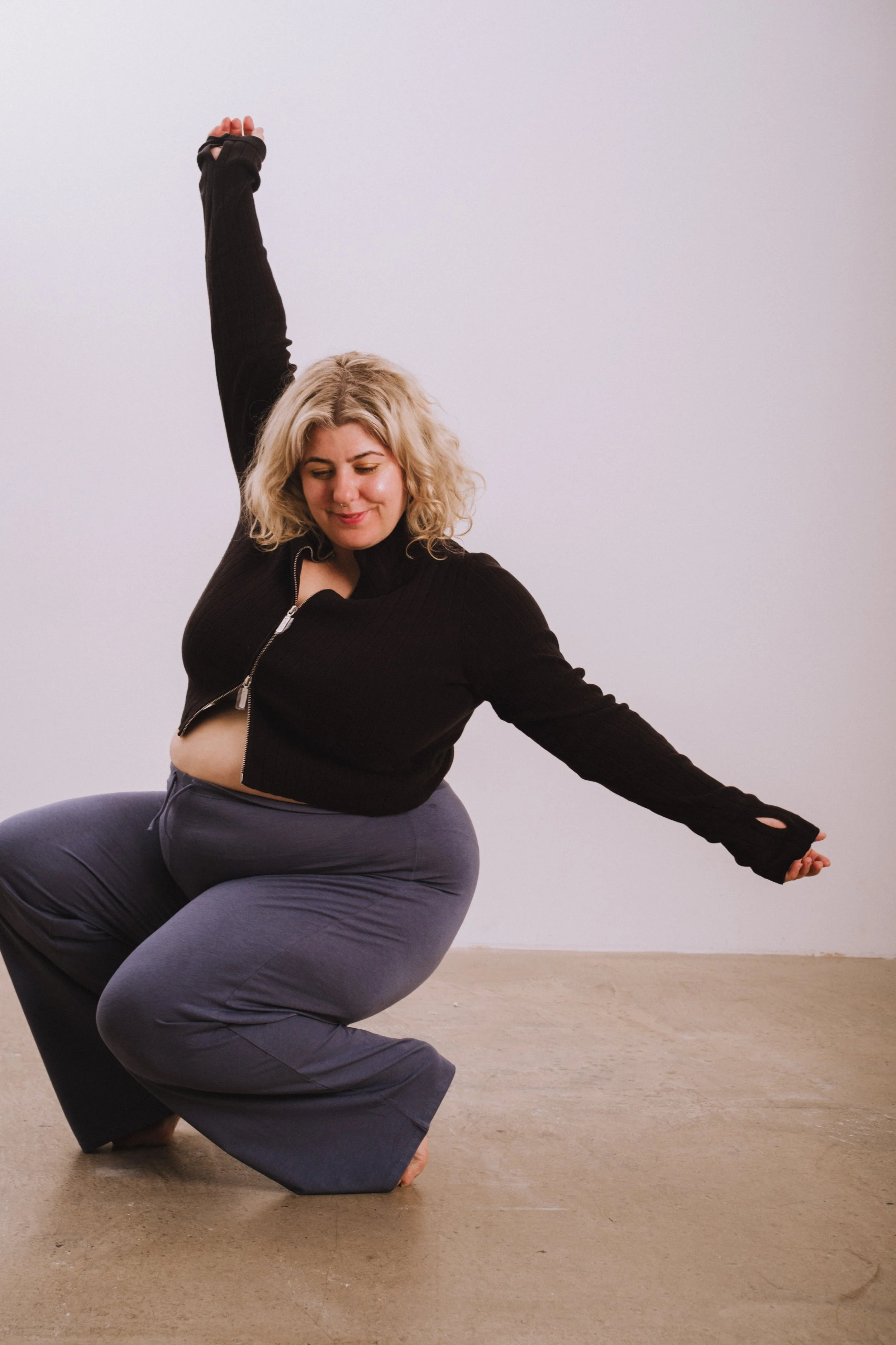 A woman with blonde curly hair doing a seated dance pose, wearing a black zip-up sweater and gray sweatpants, against a plain light-colored background.