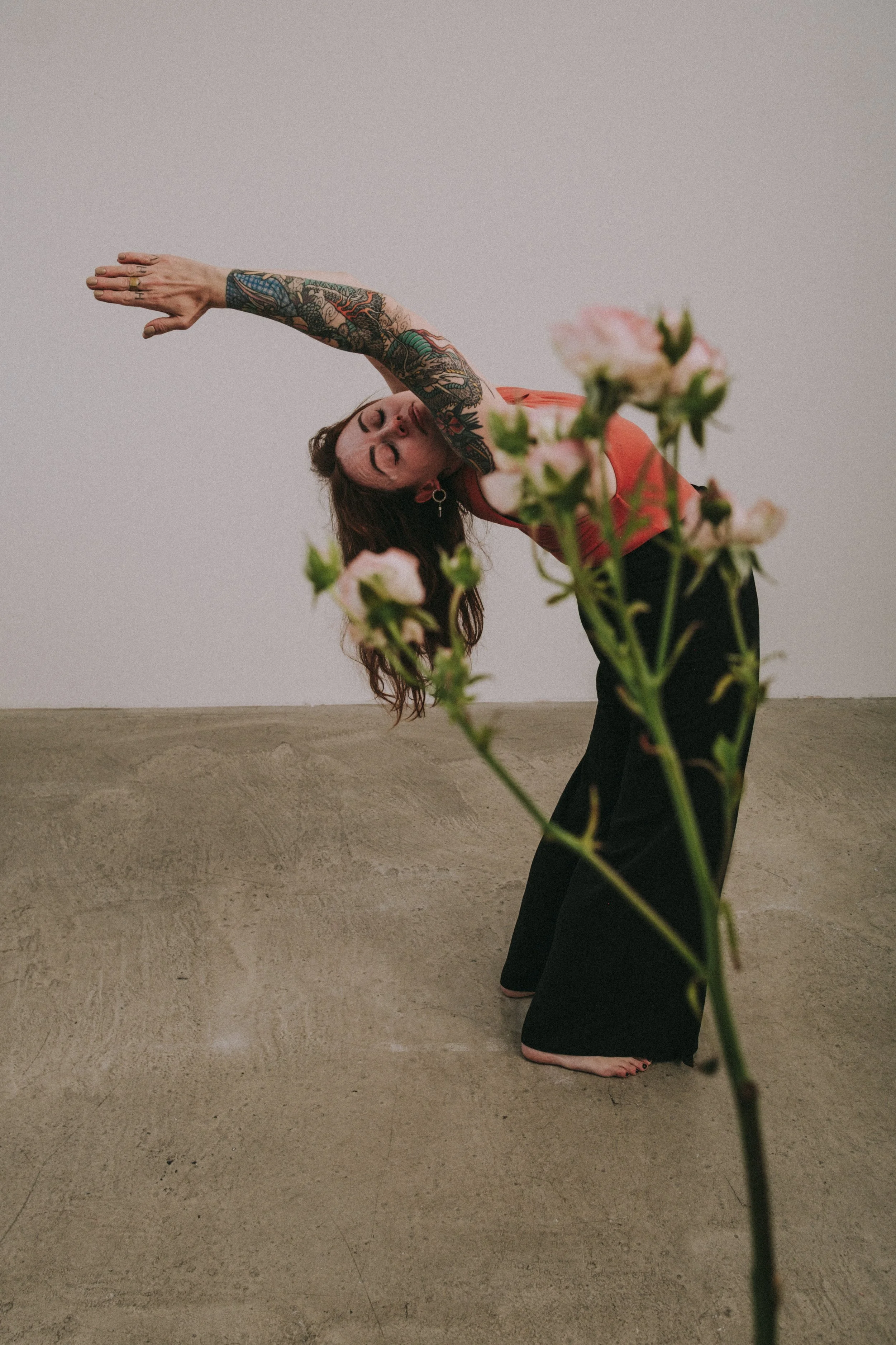 A woman with long brown hair, tattoos on her left arm, wearing a red top and black pants, performing a yoga pose with her body bent sideways and her left arm reaching over her head, near a wall. Pink flowers partially obstruct the view in the foreground.