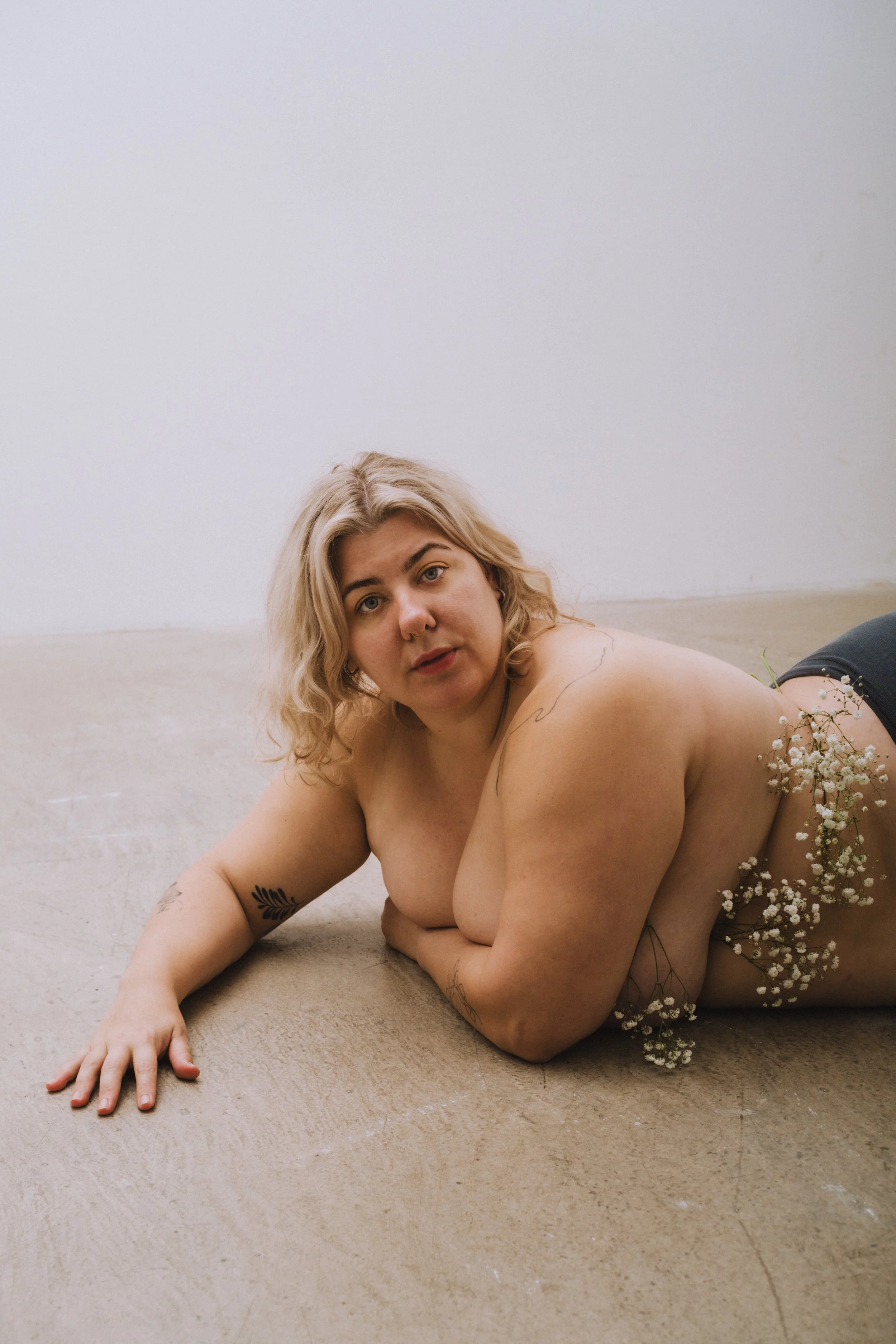 A woman with blonde hair lying on her side on a beige carpeted floor, surrounded by small white flowers, with a plain white wall in the background.