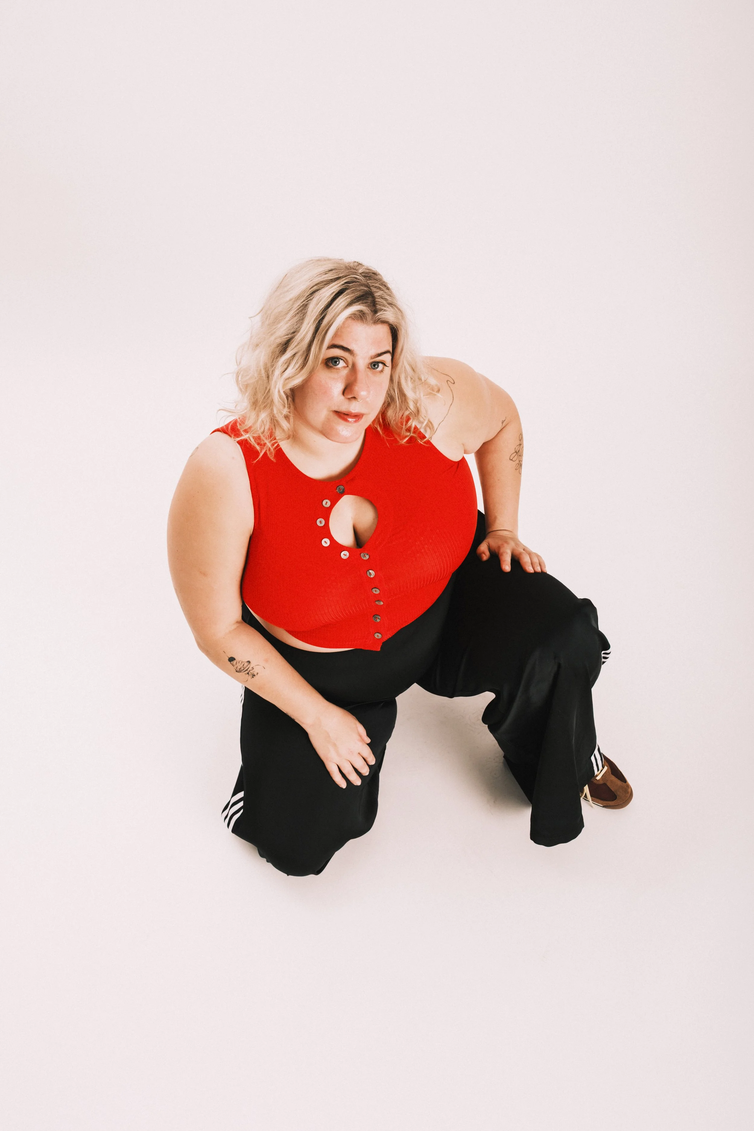 A woman with blonde curly hair wearing a red sleeveless top with a keyhole cutout, black pants with white stripes on the sides, is crouching on a white background, looking into the camera.