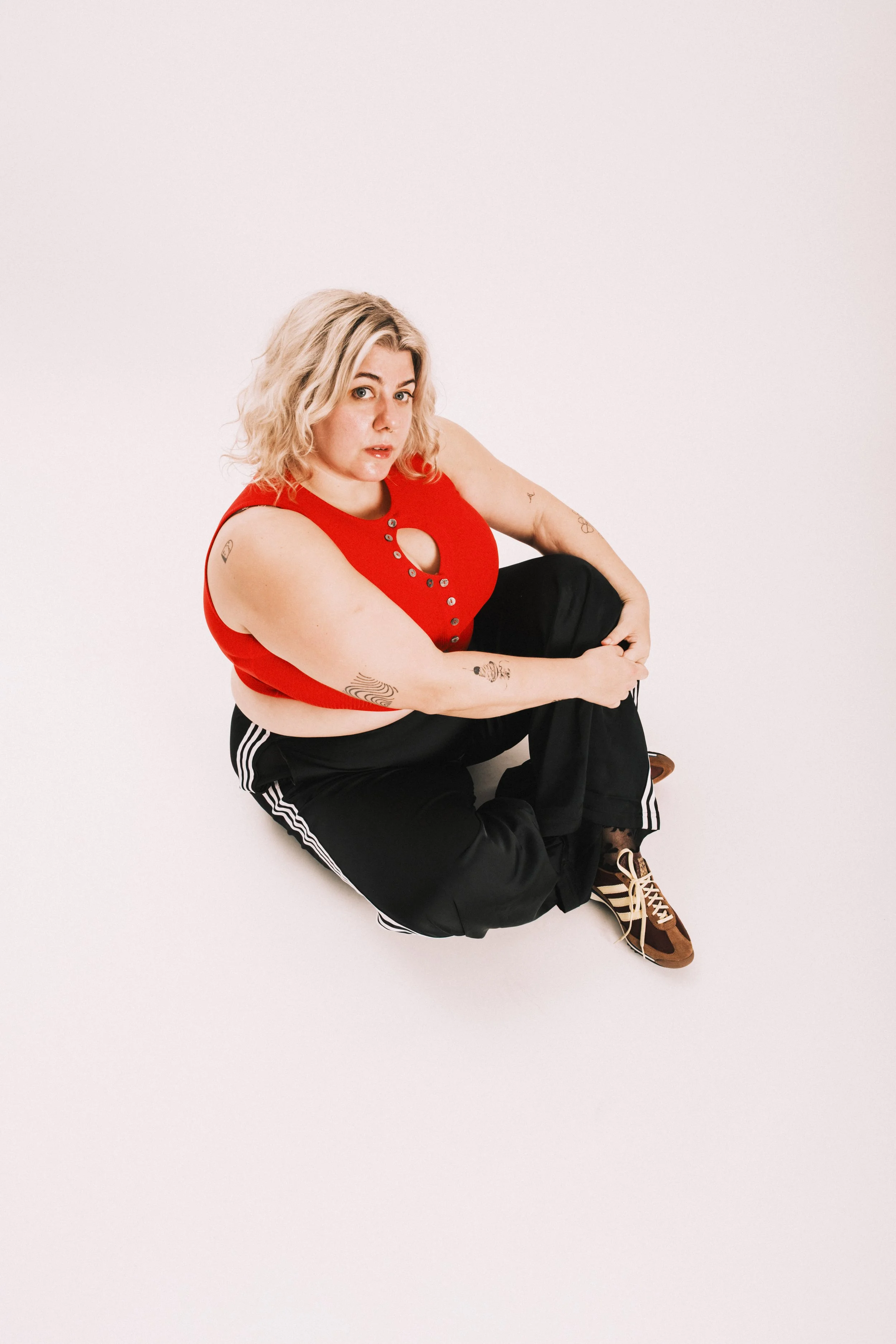 A woman with blonde curly hair, light skin, wearing a red sleeveless top with button details and black pants with white stripes, squatting against a plain light background.