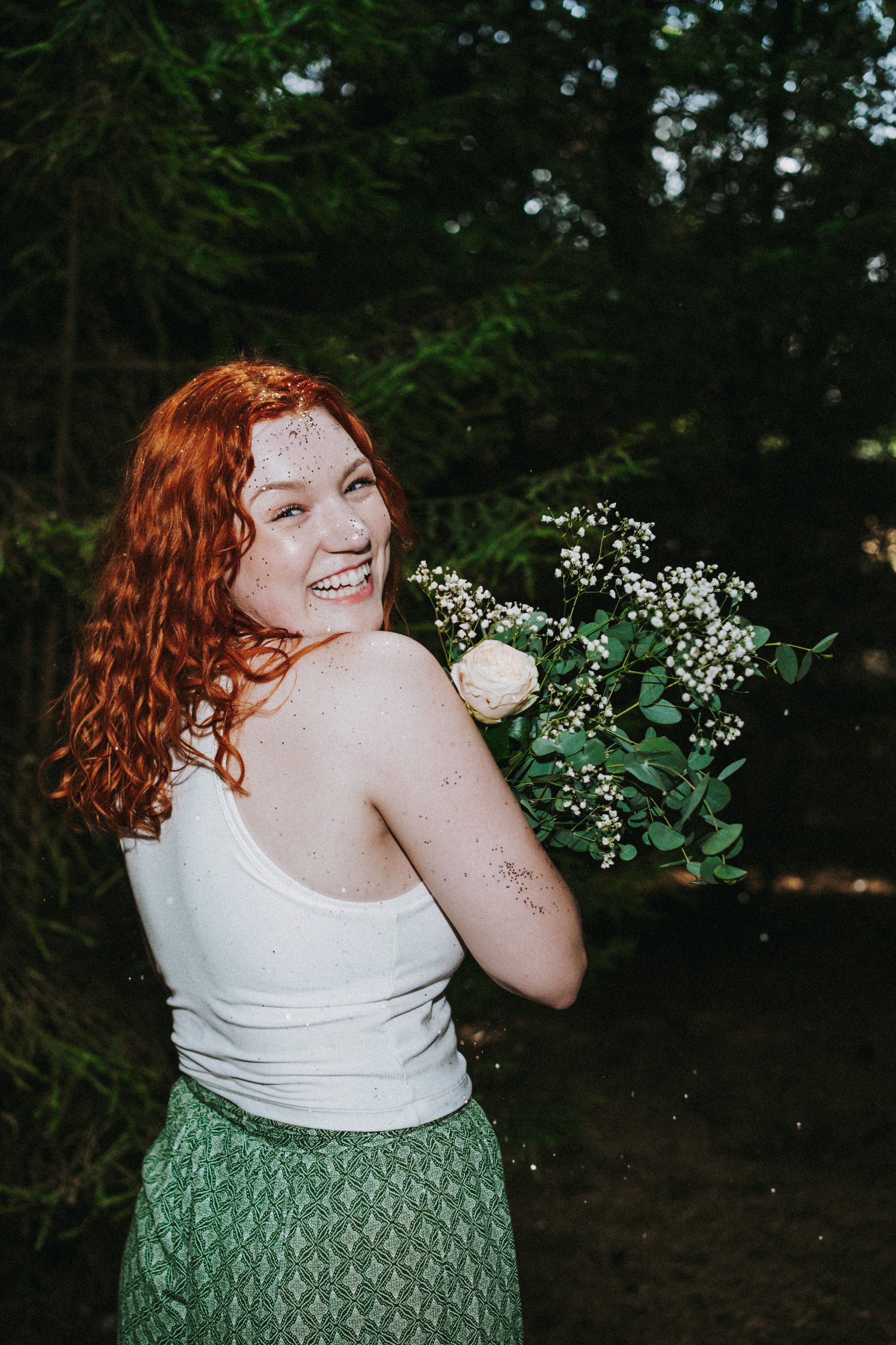 A smiling woman with red curly hair holding a bouquet of white flowers and greenery, standing outdoors in a forest setting.