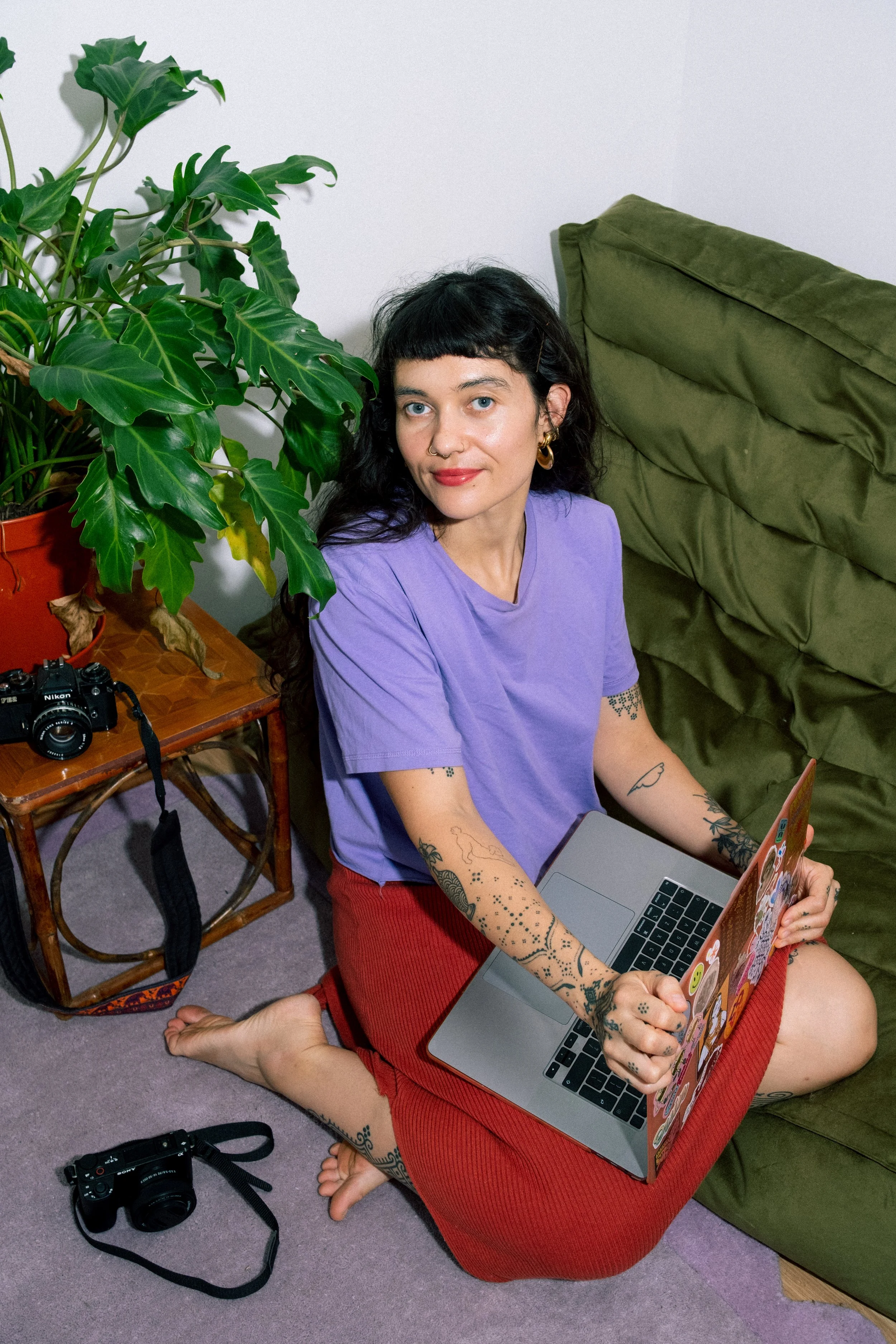 A woman with black hair and tattoos working on a laptop while sitting on the floor. She is wearing a purple T-shirt and red pants. There is a large leafy plant and a camera on a small table nearby, and a camera on the carpeted floor. She is indoors, sitting next to a green sofa.