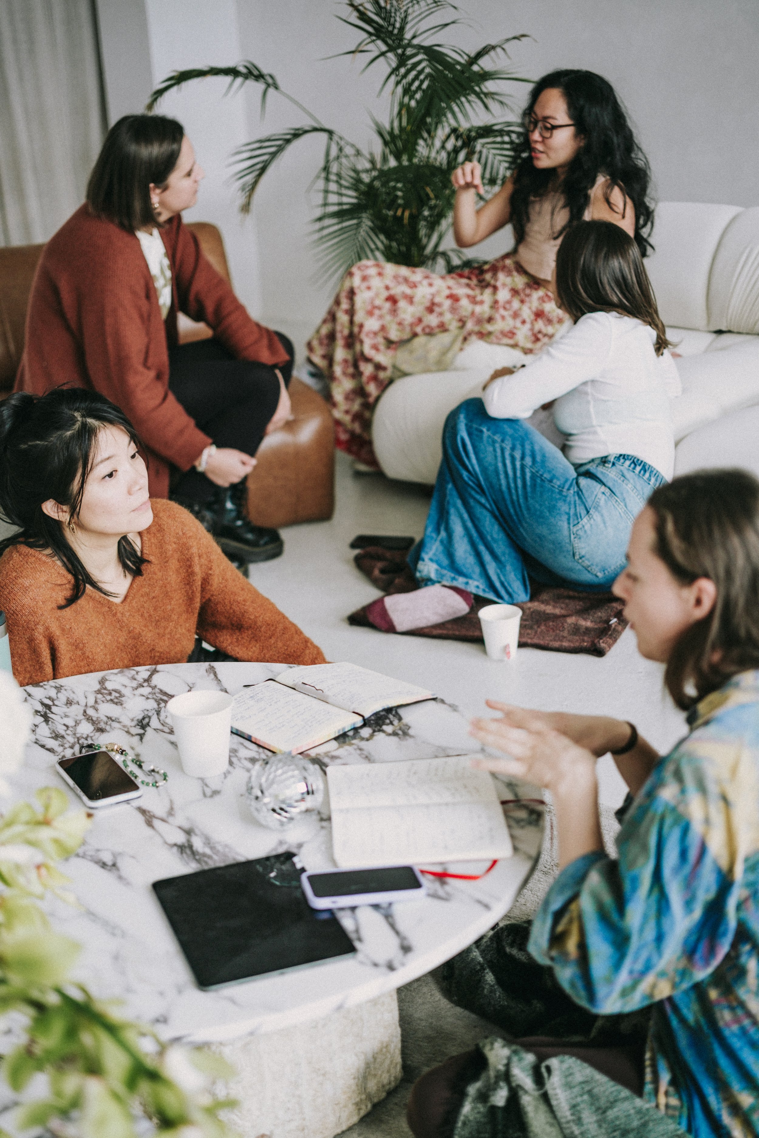 Group of women having a discussion in a cozy living room, some sitting on the couch and others around a marble table with notebooks, phones, and cups.