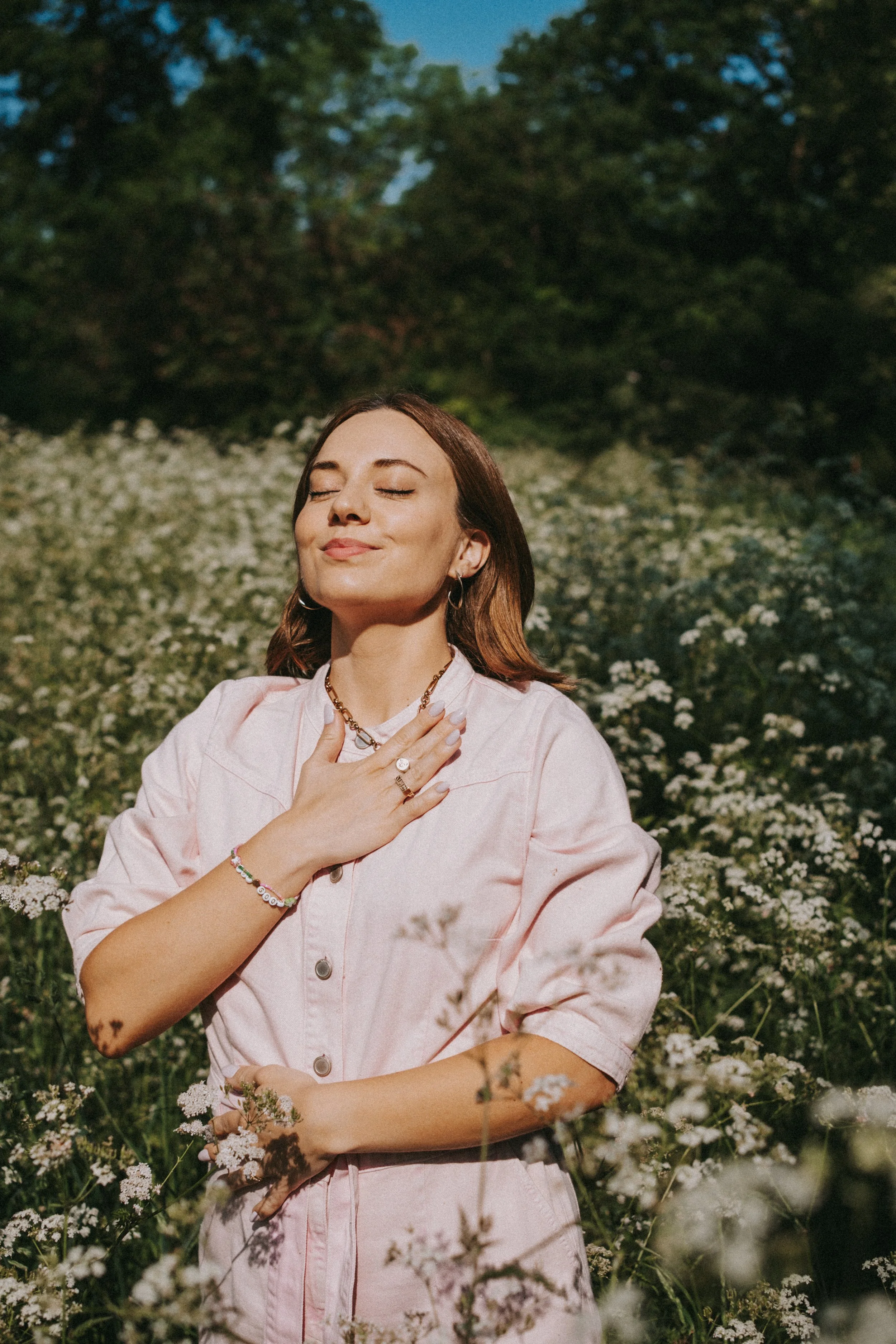 A woman with brown hair and closed eyes in a pink dress, standing in a field of small white flowers with a background of green trees and blue sky, appearing peaceful and content.