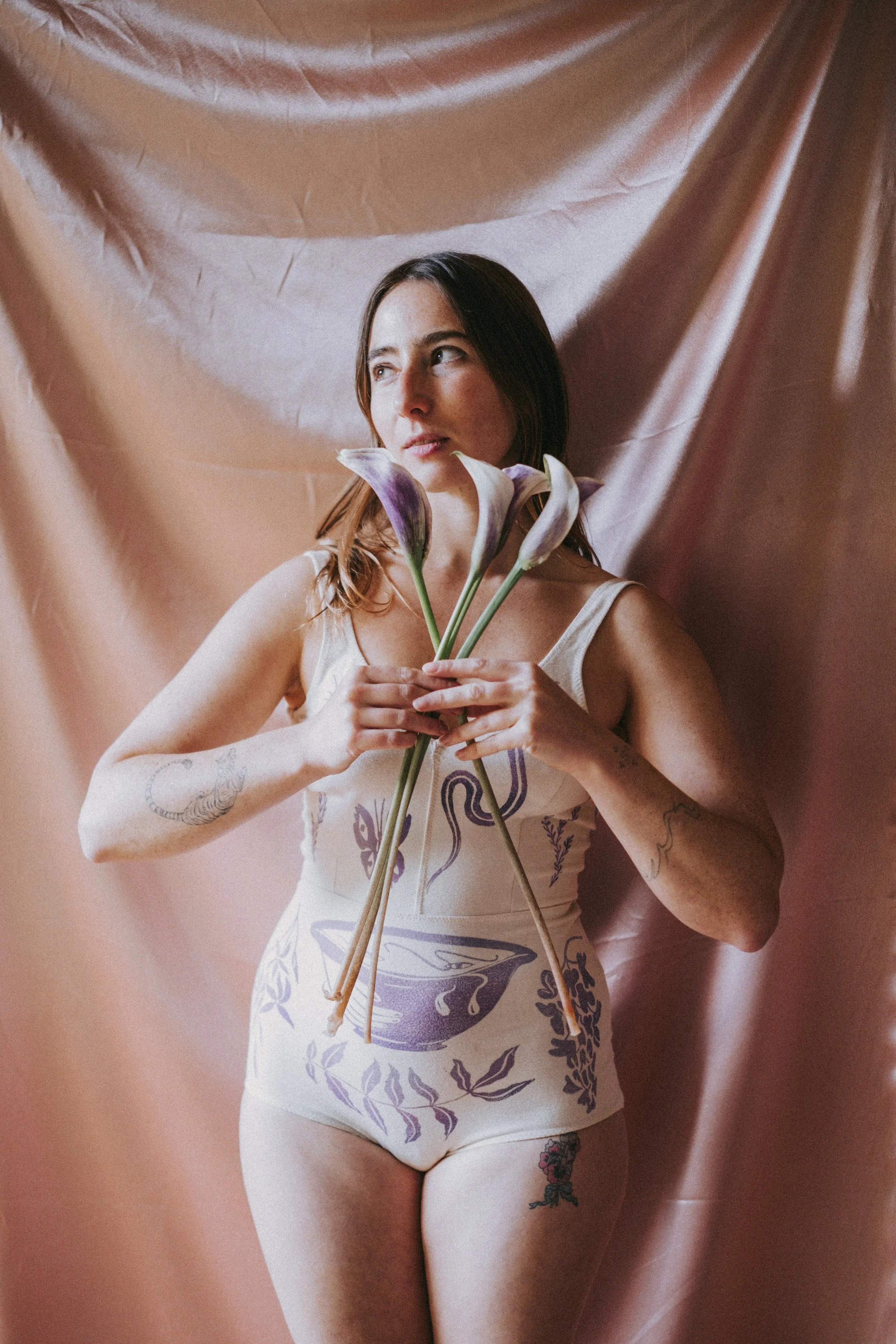 A woman with tattoos on her arm and thigh, holding three calla lilies, standing against pink satin fabric backdrop.
