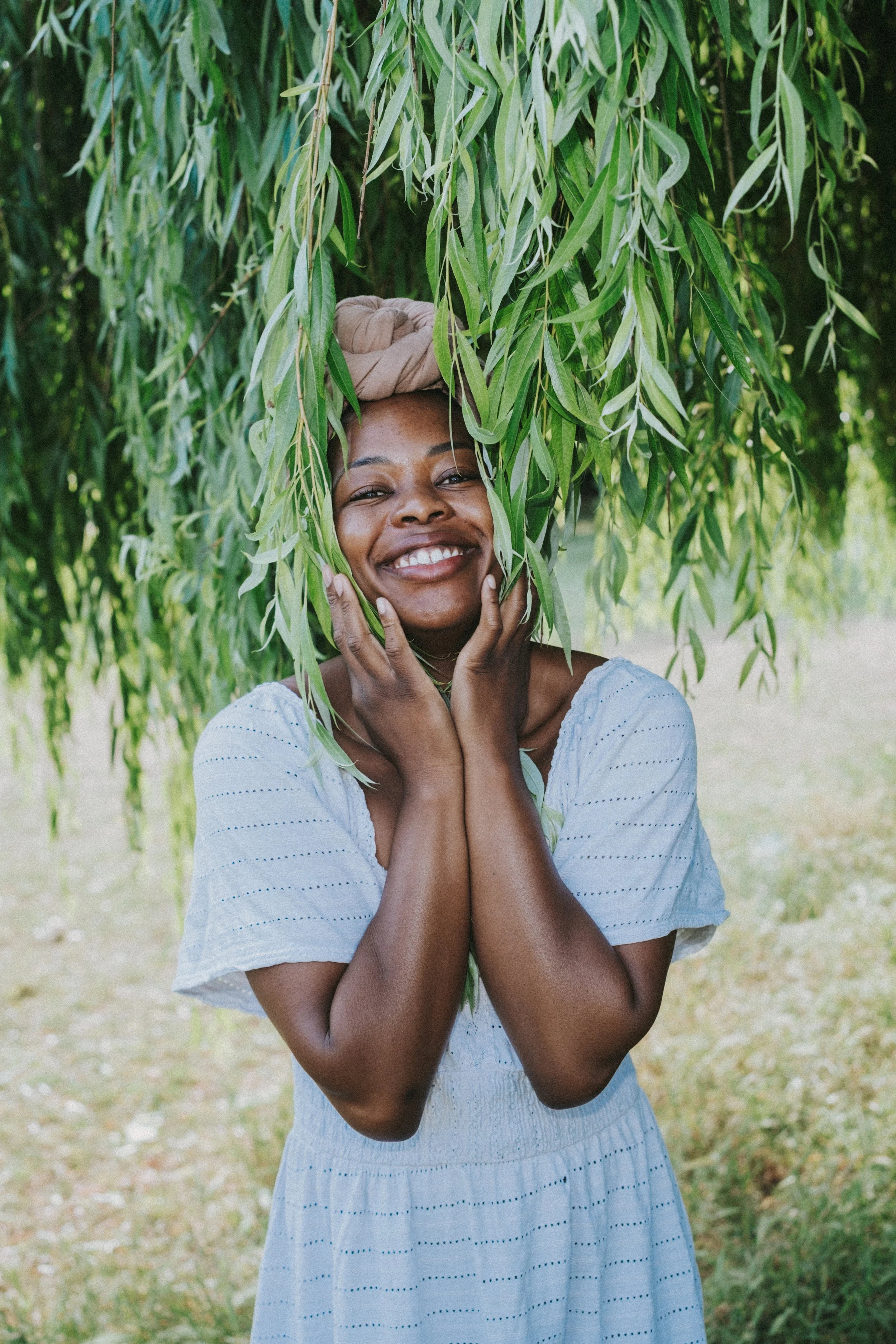 A young woman smiling with her hands on her face while standing outdoors under a leafy tree.
